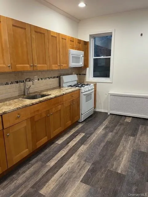 Kitchen with brown cabinetry, light stone countertops, white appliances, and radiator Kitchen with brown cabinetry, light stone countertops, white appliances, and radiator