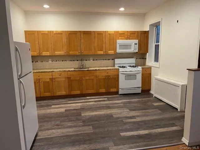 Kitchen featuring white appliances, brown cabinets, radiator heating unit, dark wood-style floors, and decorative backsplash Kitchen featuring white appliances, brown cabinets, radiator heating unit, dark wood-style floors, and decorative backsplash