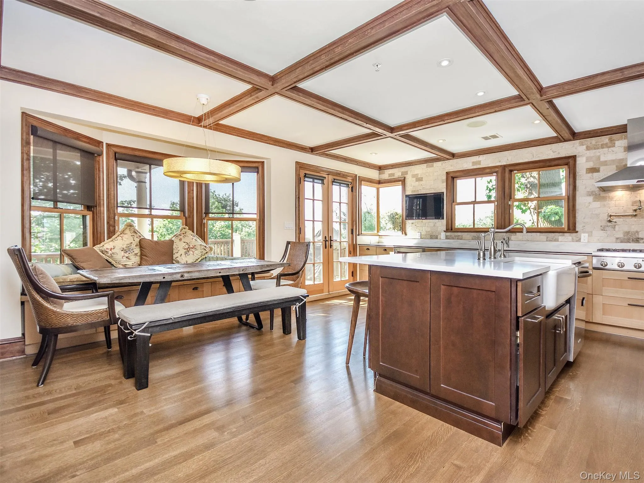 Kitchen featuring an island with sink, french doors, light wood finished floors, coffered ceiling, and a benched in dining area Kitchen featuring an island with sink, french doors, light wood finished floors, coffered ceiling, and a benched in dining area