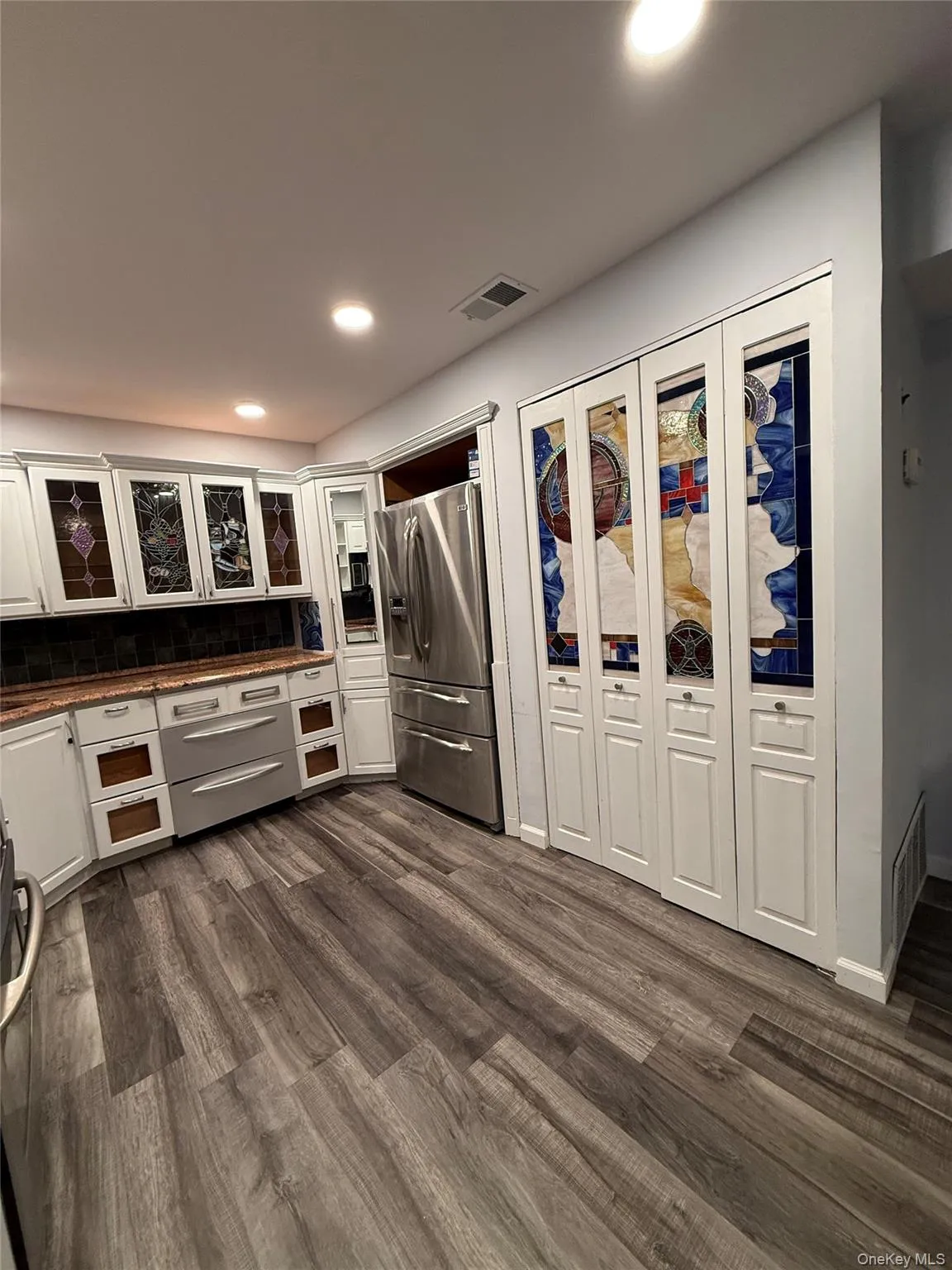 Kitchen featuring dark countertops, white cabinets, stainless steel refrigerator with ice dispenser, glass insert cabinets, and recessed lighting Kitchen featuring dark countertops, white cabinets, stainless steel refrigerator with ice dispenser, glass insert cabinets, and recessed lighting