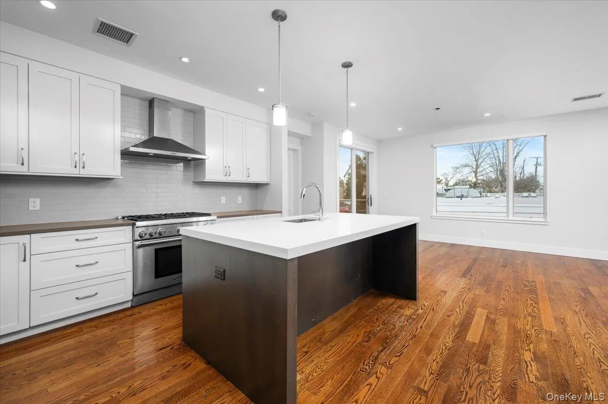Kitchen featuring high end stove, wall chimney exhaust hood, pendant lighting, tasteful backsplash, and a kitchen island with sink Kitchen featuring high end stove, wall chimney exhaust hood, pendant lighting, tasteful backsplash, and a kitchen island with sink