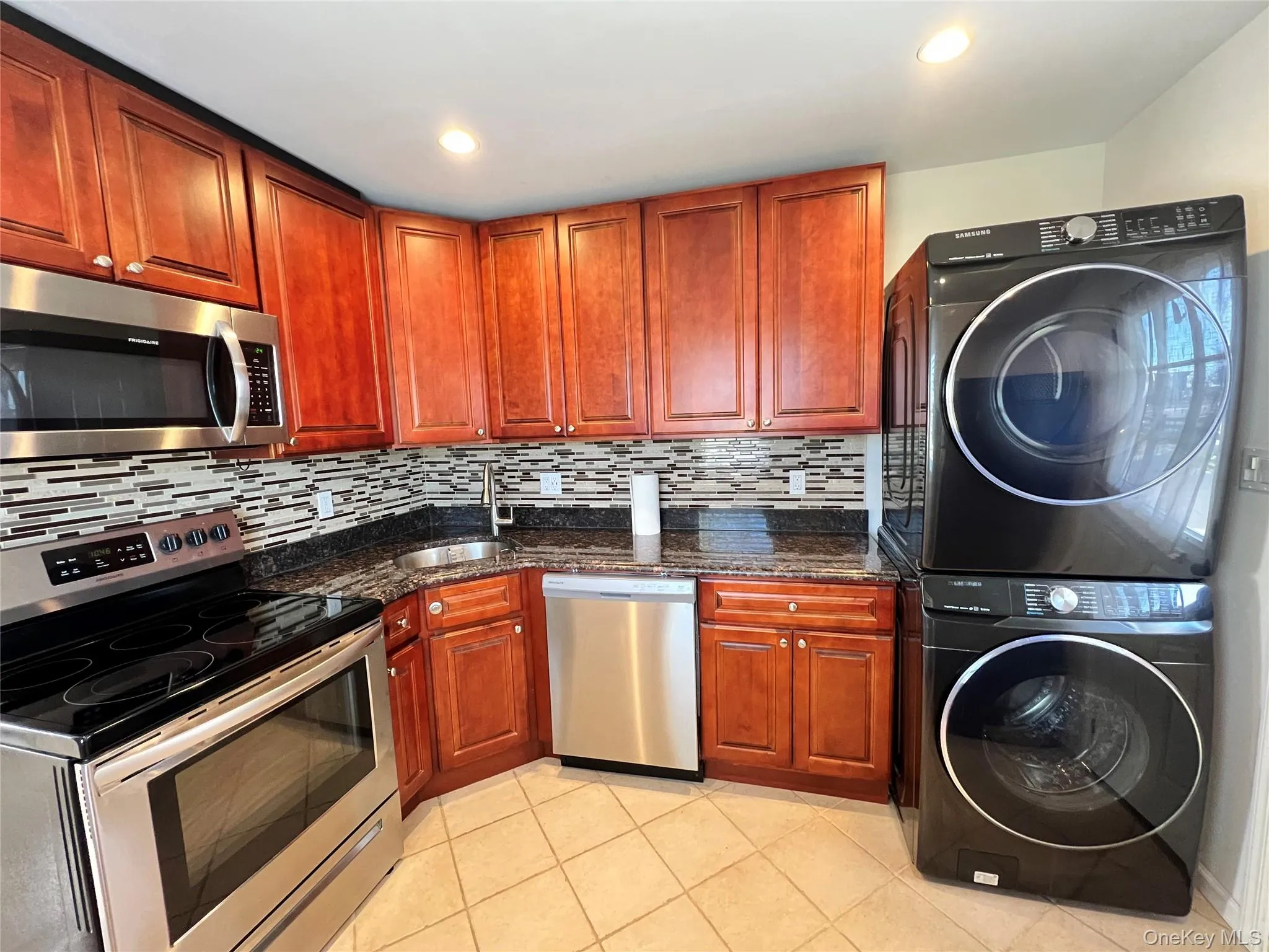 Kitchen with appliances with stainless steel finishes, dark stone countertops, stacked washer / drying machine, and light tile patterned floors Kitchen with appliances with stainless steel finishes, dark stone countertops, stacked washer / drying machine, and light tile patterned floors