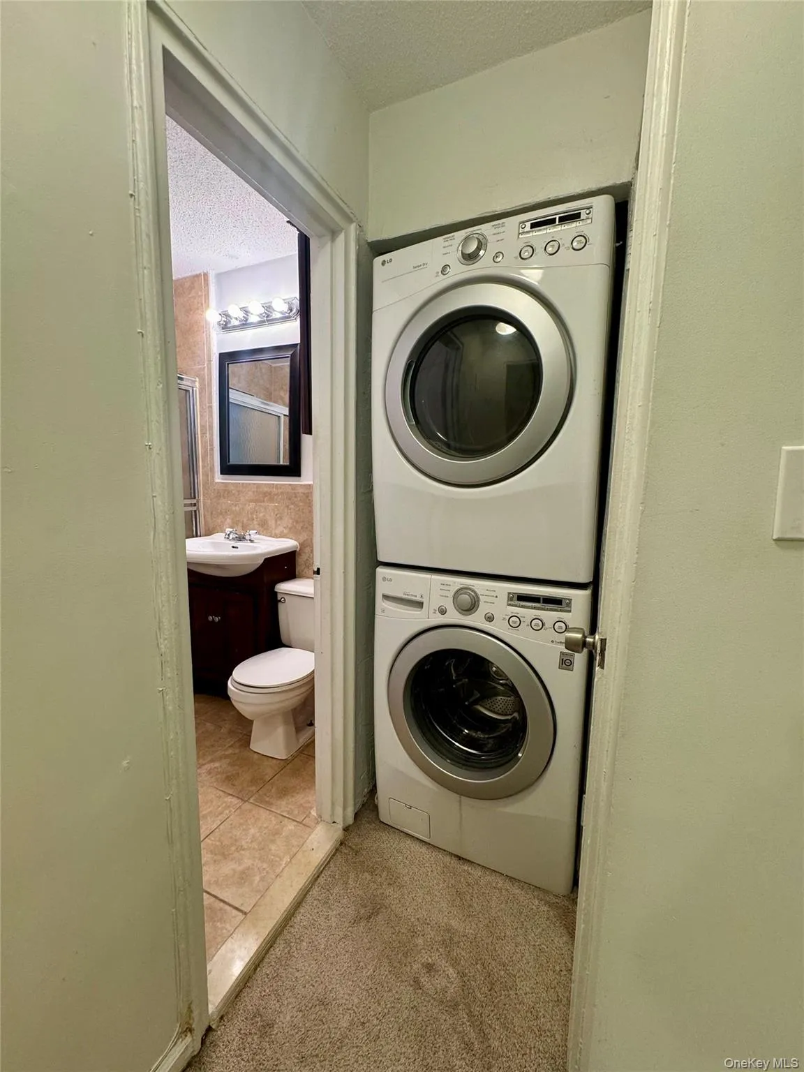 Laundry area featuring a textured ceiling, stacked washer and clothes dryer, light colored carpet, and light tile patterned floors Laundry area featuring a textured ceiling, stacked washer and clothes dryer, light colored carpet, and light tile patterned floors