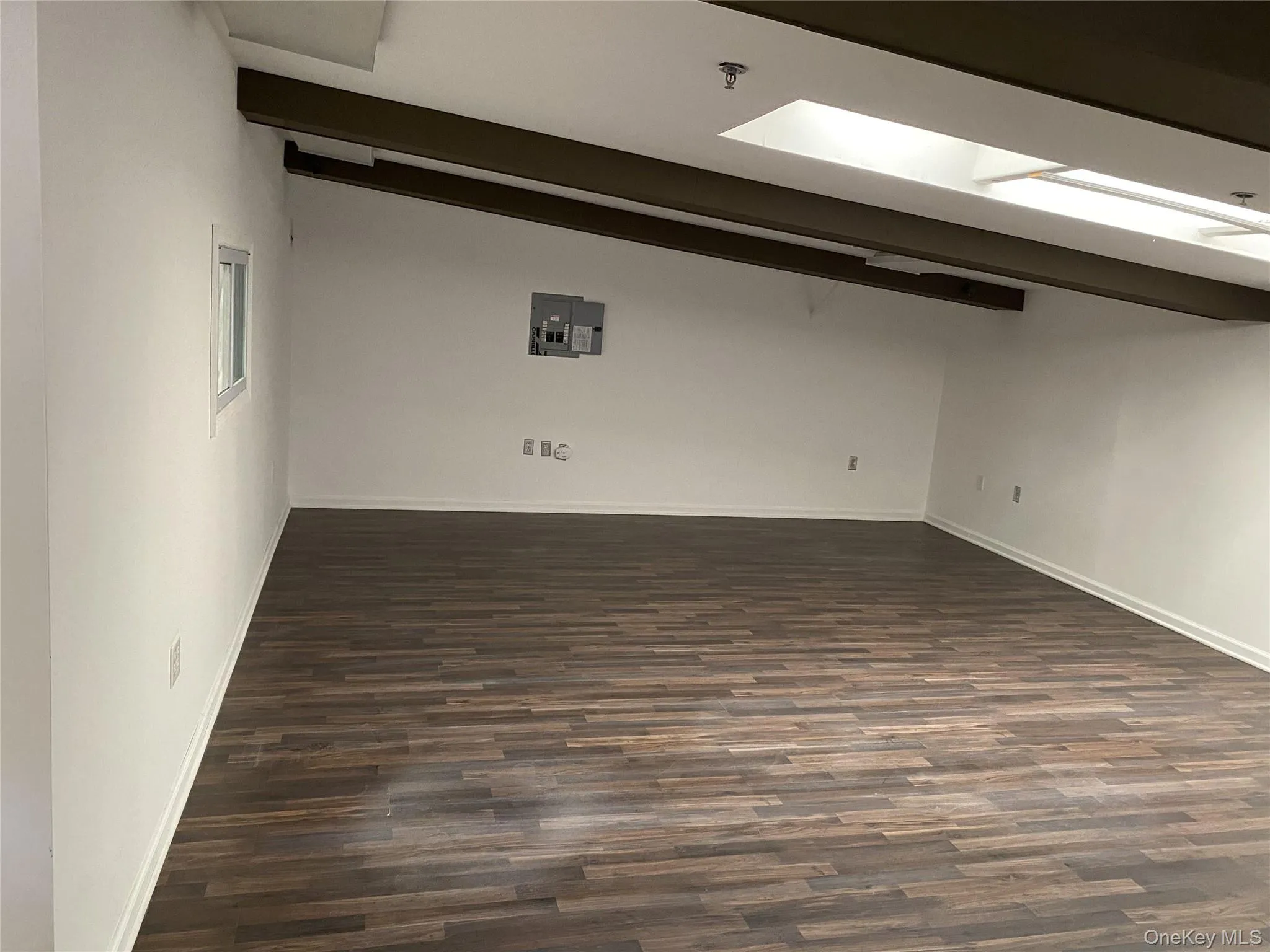 Empty room featuring dark wood-style floors, beamed ceiling, a skylight, and electric panel Empty room featuring dark wood-style floors, beamed ceiling, a skylight, and electric panel
