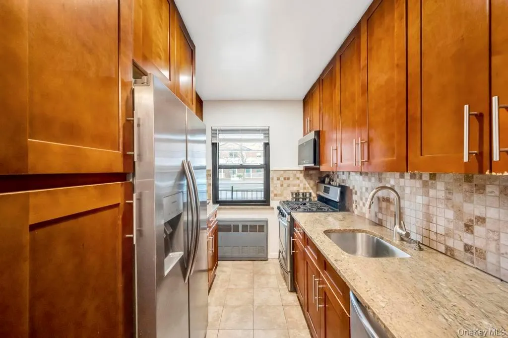 Kitchen featuring stainless steel appliances, light stone counters, brown cabinetry, decorative backsplash, and light tile patterned floors Kitchen featuring stainless steel appliances, light stone counters, brown cabinetry, decorative backsplash, and light tile patterned floors