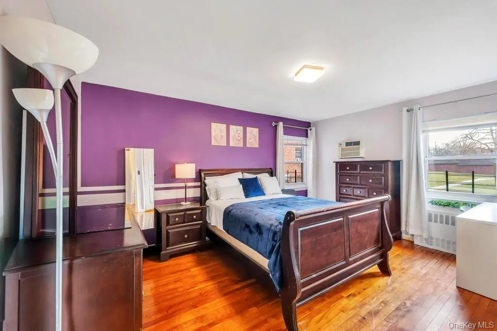 Bedroom featuring multiple windows, wood-type flooring, a wall unit AC, and radiator heating unit Bedroom featuring multiple windows, wood-type flooring, a wall unit AC, and radiator heating unit