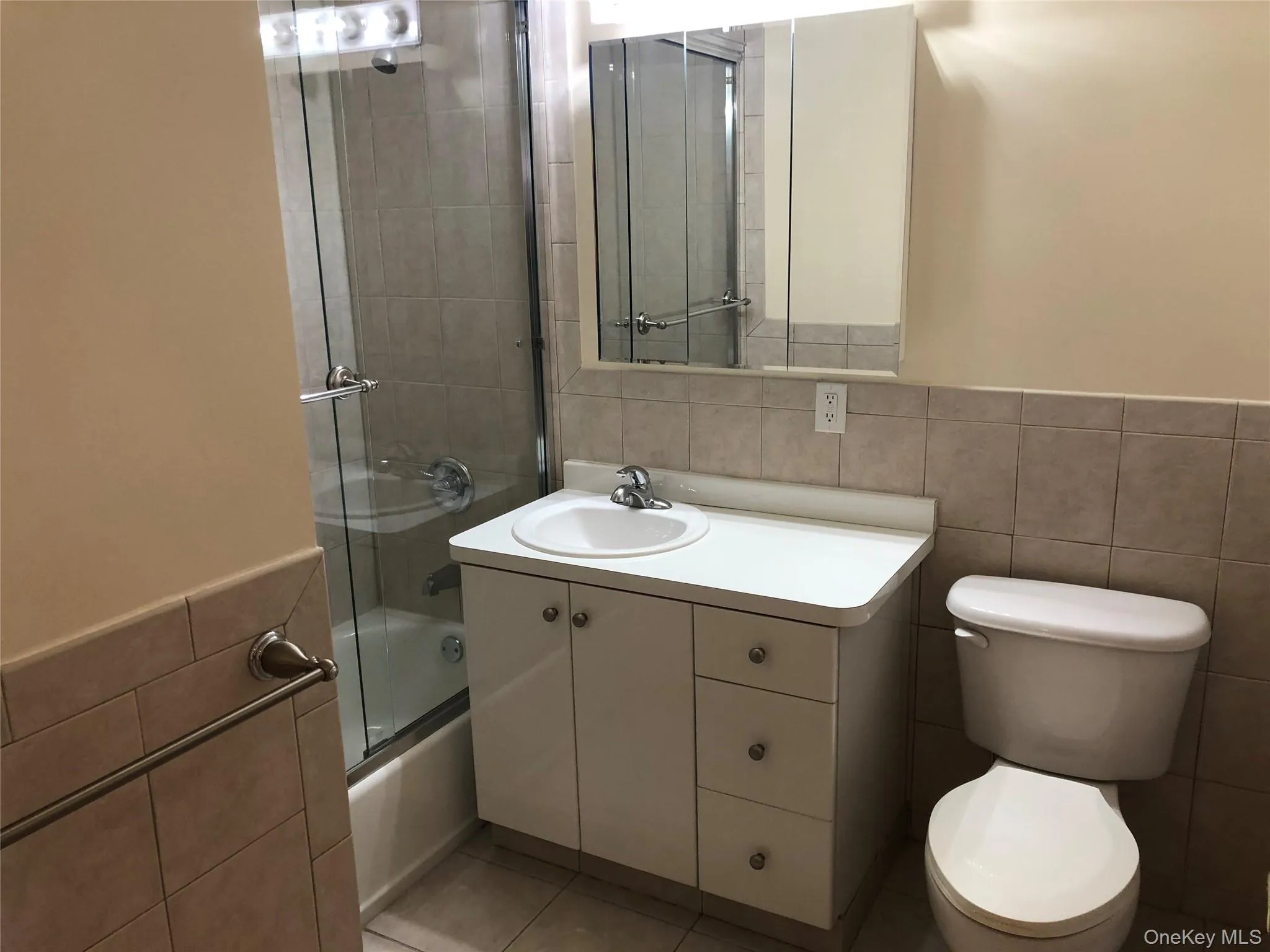 Full bathroom with tile walls, shower / bath combination with glass door, wainscoting, vanity, and light tile patterned floors Full bathroom with tile walls, shower / bath combination with glass door, wainscoting, vanity, and light tile patterned floors