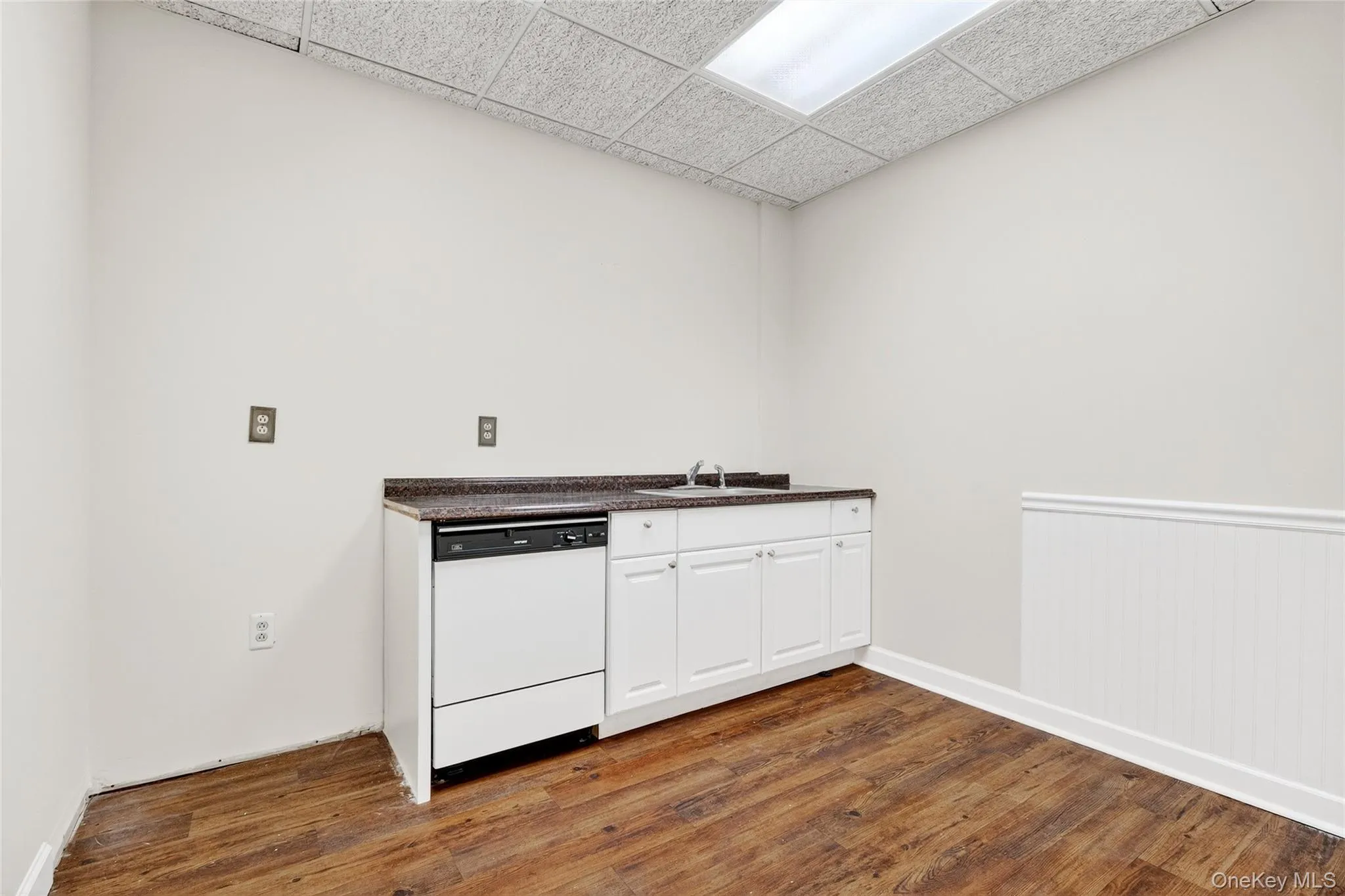 Kitchen featuring a drop ceiling, dark countertops, dishwasher, white cabinetry, and dark wood-type flooring Kitchen featuring a drop ceiling, dark countertops, dishwasher, white cabinetry, and dark wood-type flooring