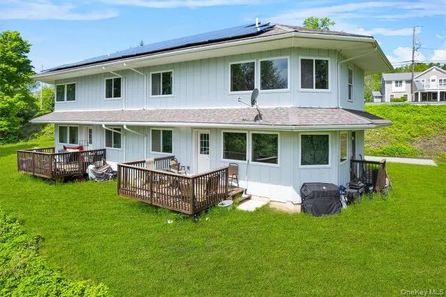 Rear view of house featuring roof mounted solar panels, a wooden deck, a lawn, and board and batten siding Rear view of house featuring roof mounted solar panels, a wooden deck, a lawn, and board and batten siding