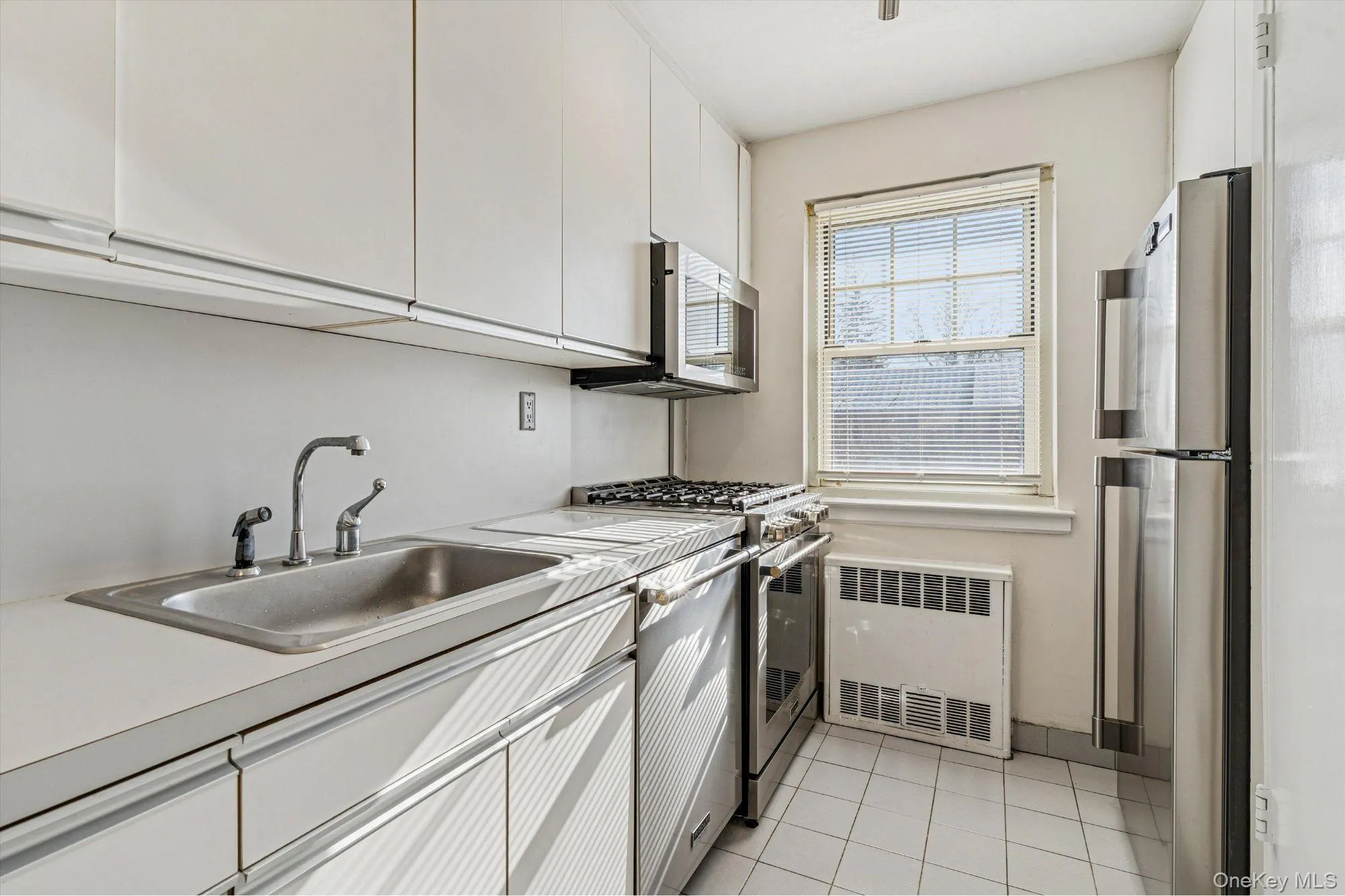 Kitchen with white cabinetry, appliances with stainless steel finishes, radiator heating unit, light countertops, and light tile patterned floors Kitchen with white cabinetry, appliances with stainless steel finishes, radiator heating unit, light countertops, and light tile patterned floors