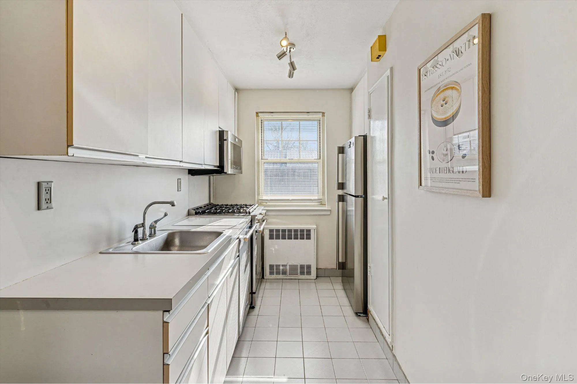 Kitchen featuring appliances with stainless steel finishes, light countertops, white cabinets, radiator heating unit, and light tile patterned flooring Kitchen featuring appliances with stainless steel finishes, light countertops, white cabinets, radiator heating unit, and light tile patterned flooring