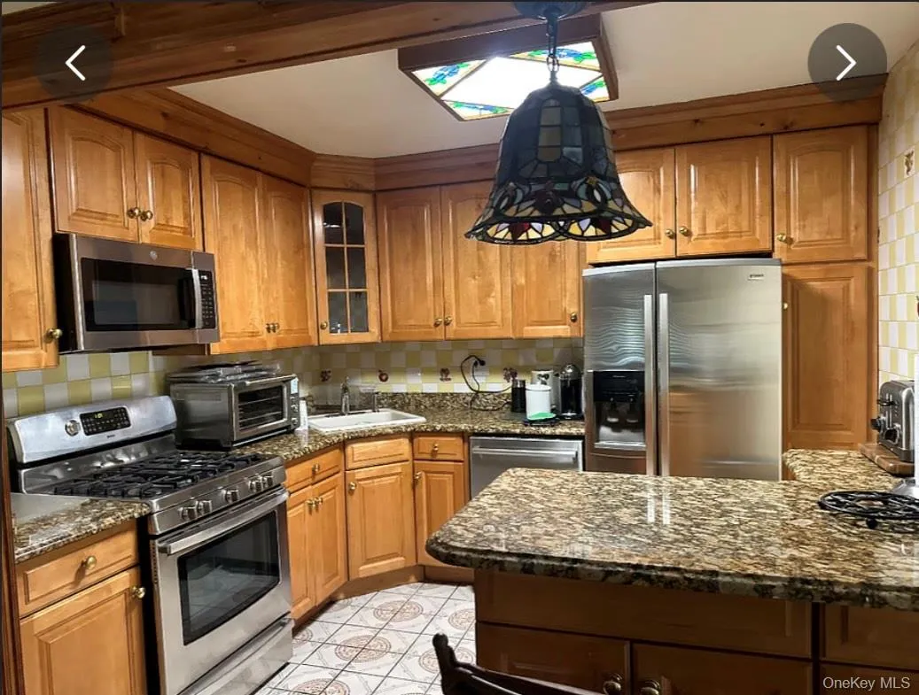 Kitchen with stainless steel appliances, brown cabinetry, dark stone countertops, and backsplash Kitchen with stainless steel appliances, brown cabinetry, dark stone countertops, and backsplash