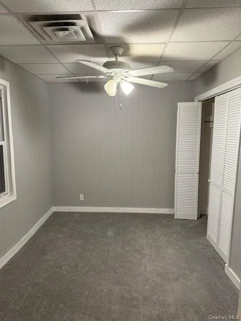 Unfurnished bedroom with wooden walls, a drop ceiling, dark colored carpet, a closet, and a ceiling fan Unfurnished bedroom with wooden walls, a drop ceiling, dark colored carpet, a closet, and a ceiling fan