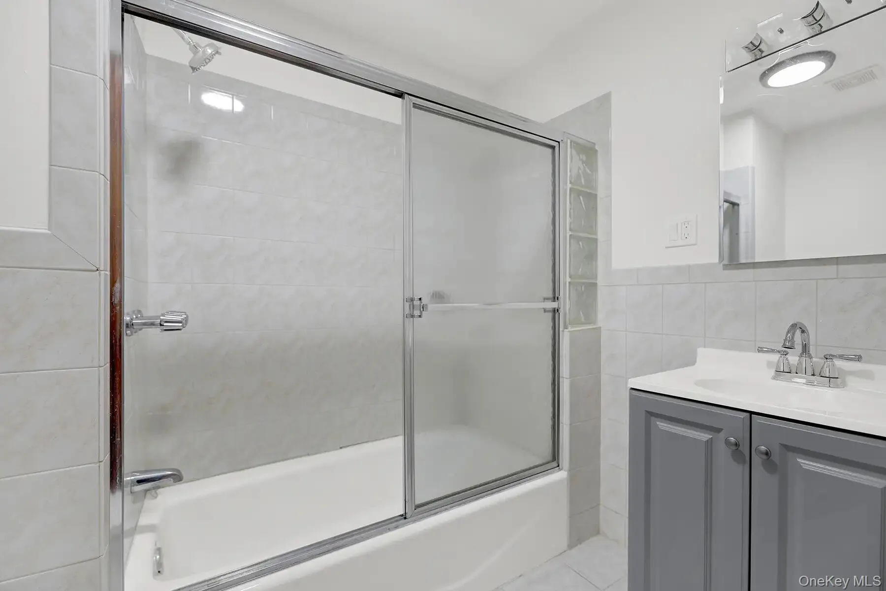 Bathroom featuring a tub and shower combination with sliding glass doors, a vanity with a white countertop and gray cabinetry, a wall-mounted mirror, and tile flooring Bathroom featuring a tub and shower combination with sliding glass doors, a vanity with a white countertop and gray cabinetry, a wall-mounted mirror, and tile flooring