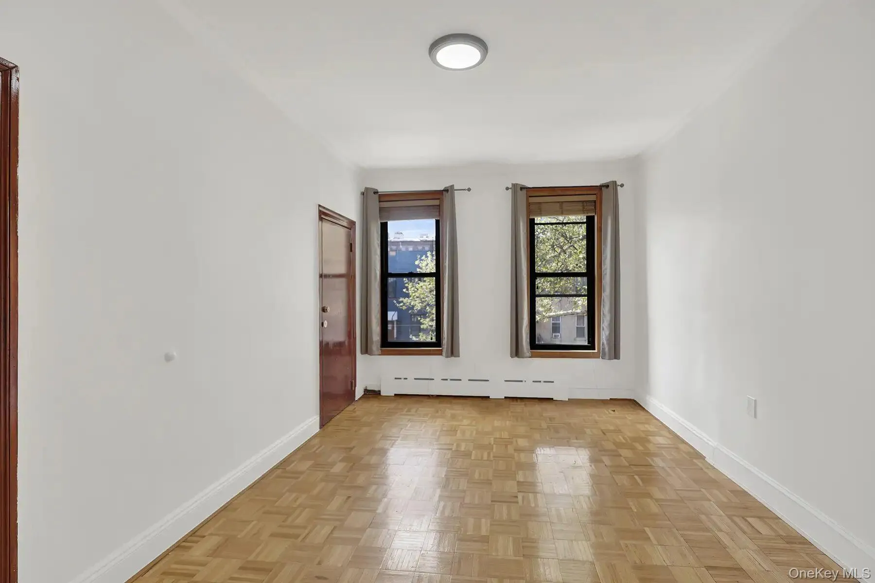 Spacious room featuring light wood-finish parquet flooring, two large windows with dark frames, and white painted walls Spacious room featuring light wood-finish parquet flooring, two large windows with dark frames, and white painted walls
