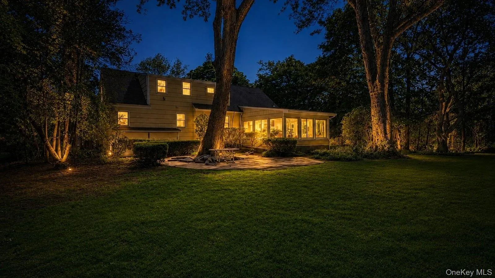 real estate listing photo showing the rear exterior, backyard lawn, patio area, mature trees, and Florida room at 9 Sheep Pasture Lane in Huntington, NY 11743 real estate listing photo showing the rear exterior, backyard lawn, patio area, mature trees, and Florida room at 9 Sheep Pasture Lane in Huntington, NY 11743