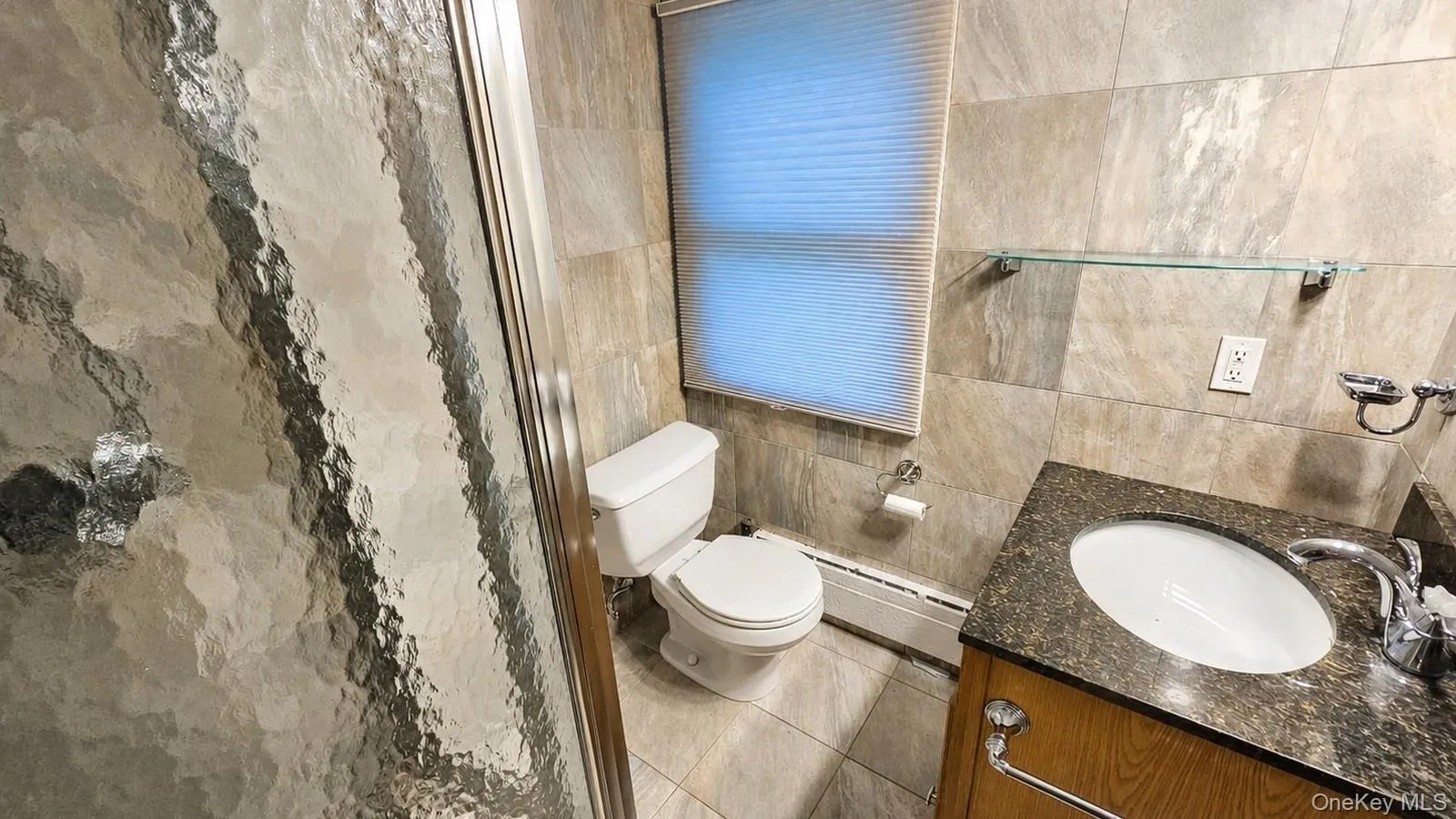 Bathroom featuring a glass shower enclosure, a vanity with a dark countertop and undermount sink, a white toilet, and neutral-toned tiled walls and flooring Bathroom featuring a glass shower enclosure, a vanity with a dark countertop and undermount sink, a white toilet, and neutral-toned tiled walls and flooring