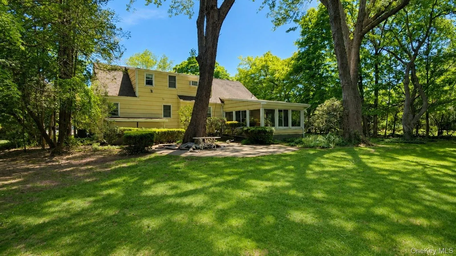 real estate listing photo showing the rear exterior, backyard lawn, patio area, mature trees, and Florida room at 9 Sheep Pasture Lane in Huntington, NY 11743 real estate listing photo showing the rear exterior, backyard lawn, patio area, mature trees, and Florida room at 9 Sheep Pasture Lane in Huntington, NY 11743