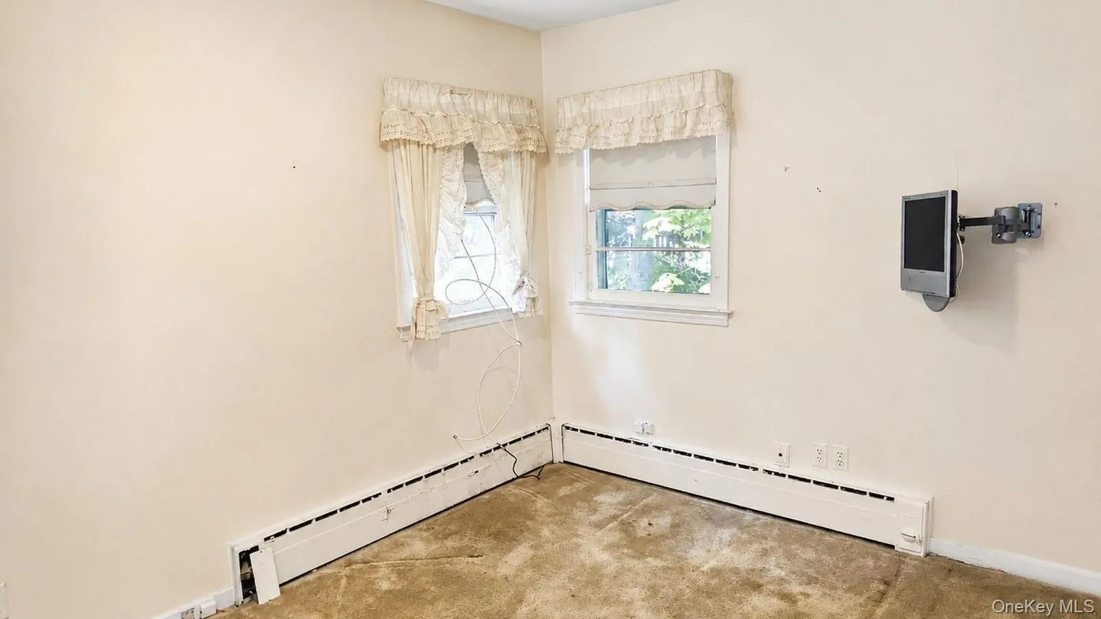 real estate listing photo of a bedroom at 9 Sheep Pasture Lane in Huntington, NY, 11743, showing two windows, wall-mounted TV, baseboard heating, and visible carpet condition. real estate listing photo of a bedroom at 9 Sheep Pasture Lane in Huntington, NY, 11743, showing two windows, wall-mounted TV, baseboard heating, and visible carpet condition.