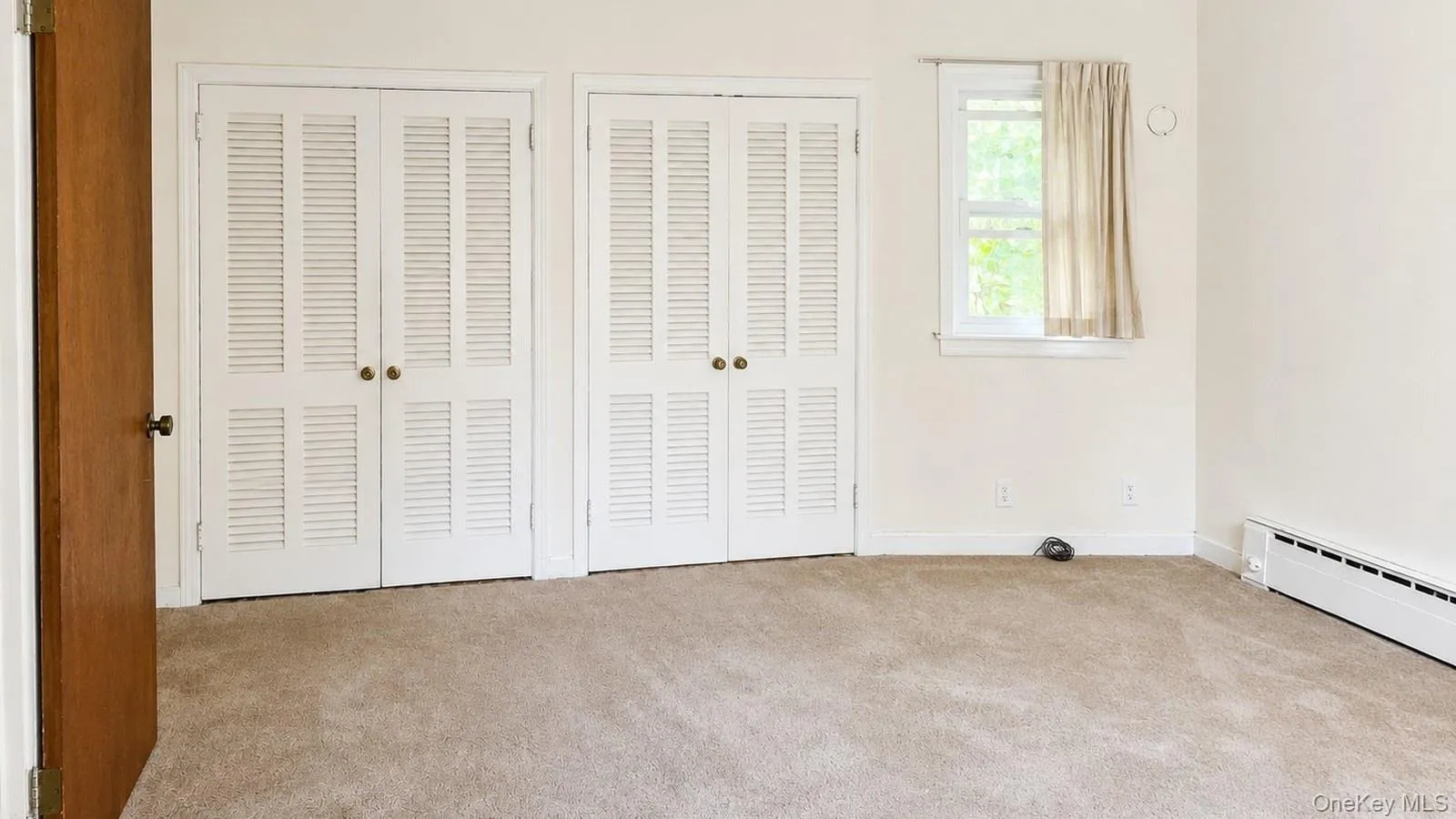 real estate listing photo showing the primary bedroom at 9 Sheep Pasture Lane in Huntington, NY 11743 with two closet doors, window, baseboard heating, and visible carpet condition. real estate listing photo showing the primary bedroom at 9 Sheep Pasture Lane in Huntington, NY 11743 with two closet doors, window, baseboard heating, and visible carpet condition.