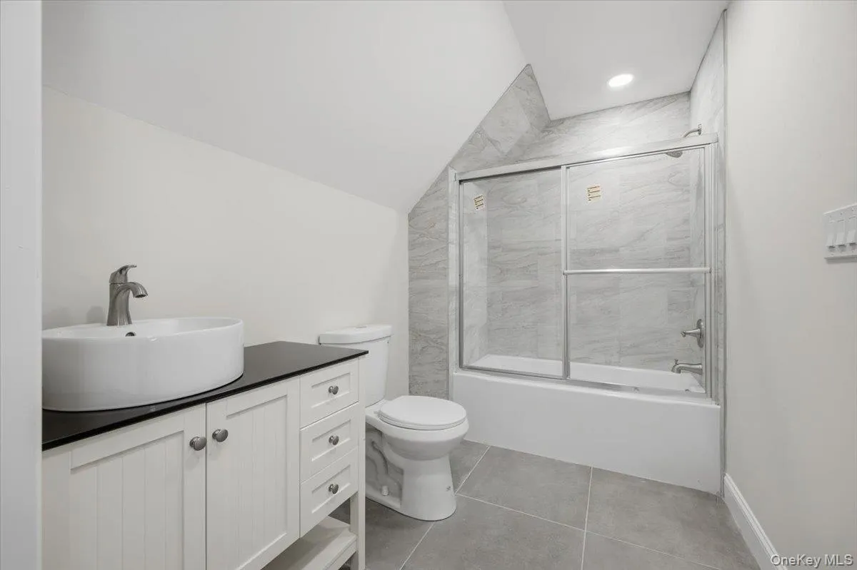 Modern bathroom with a white vessel sink on a dark countertop, white vanity cabinetry with brushed nickel hardware, a white toilet, and a tiled shower/tub combination with a sliding glass door Modern bathroom with a white vessel sink on a dark countertop, white vanity cabinetry with brushed nickel hardware, a white toilet, and a tiled shower/tub combination with a sliding glass door