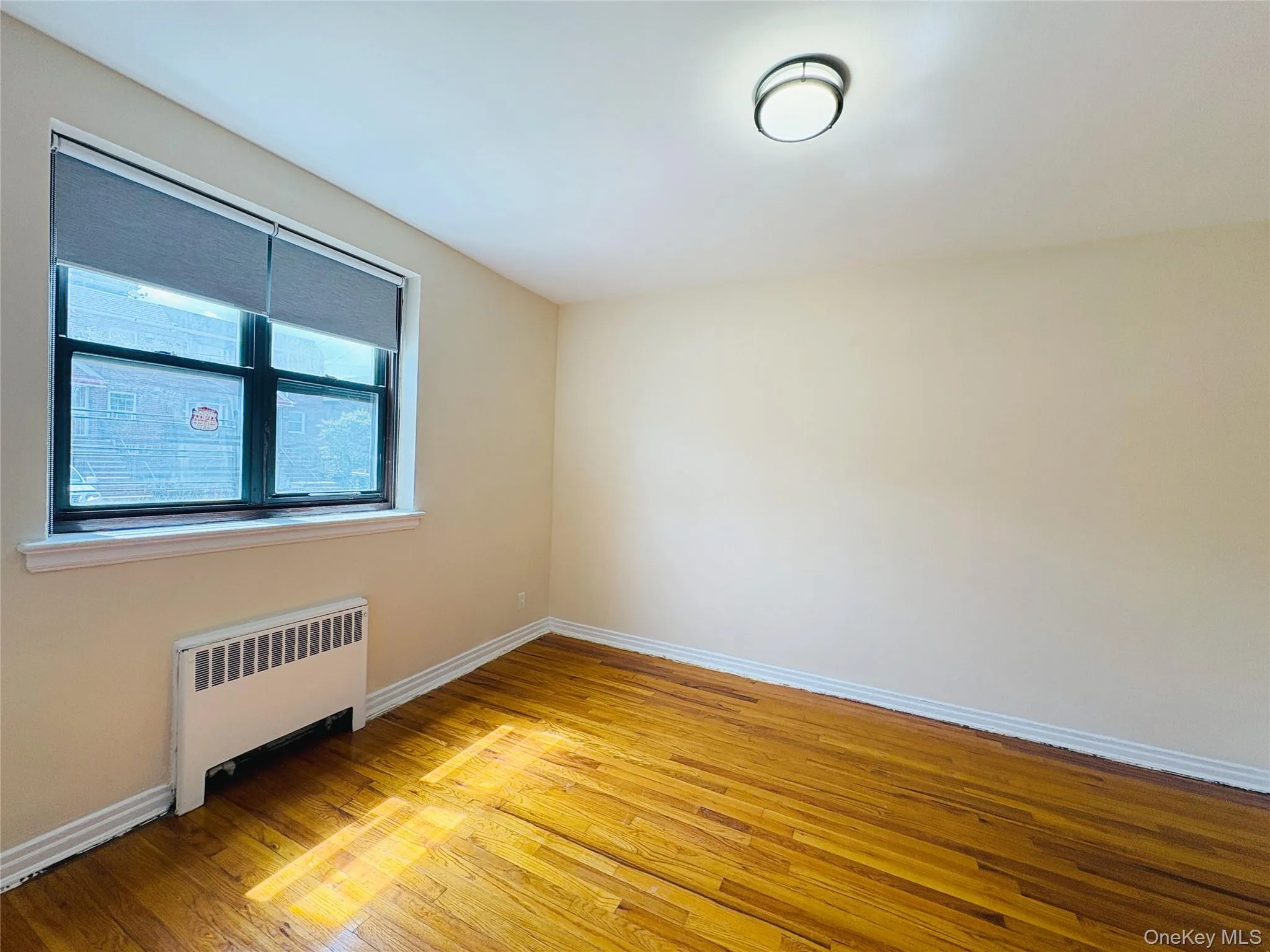 Room featuring wood-finish flooring, neutral wall tones, a large window with roller shades, and a ceiling-mounted light fixture Room featuring wood-finish flooring, neutral wall tones, a large window with roller shades, and a ceiling-mounted light fixture