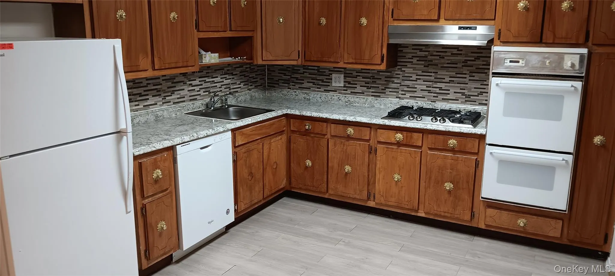 Kitchen with white appliances, light countertops, brown cabinets, tasteful backsplash, and under cabinet range hood Kitchen with white appliances, light countertops, brown cabinets, tasteful backsplash, and under cabinet range hood