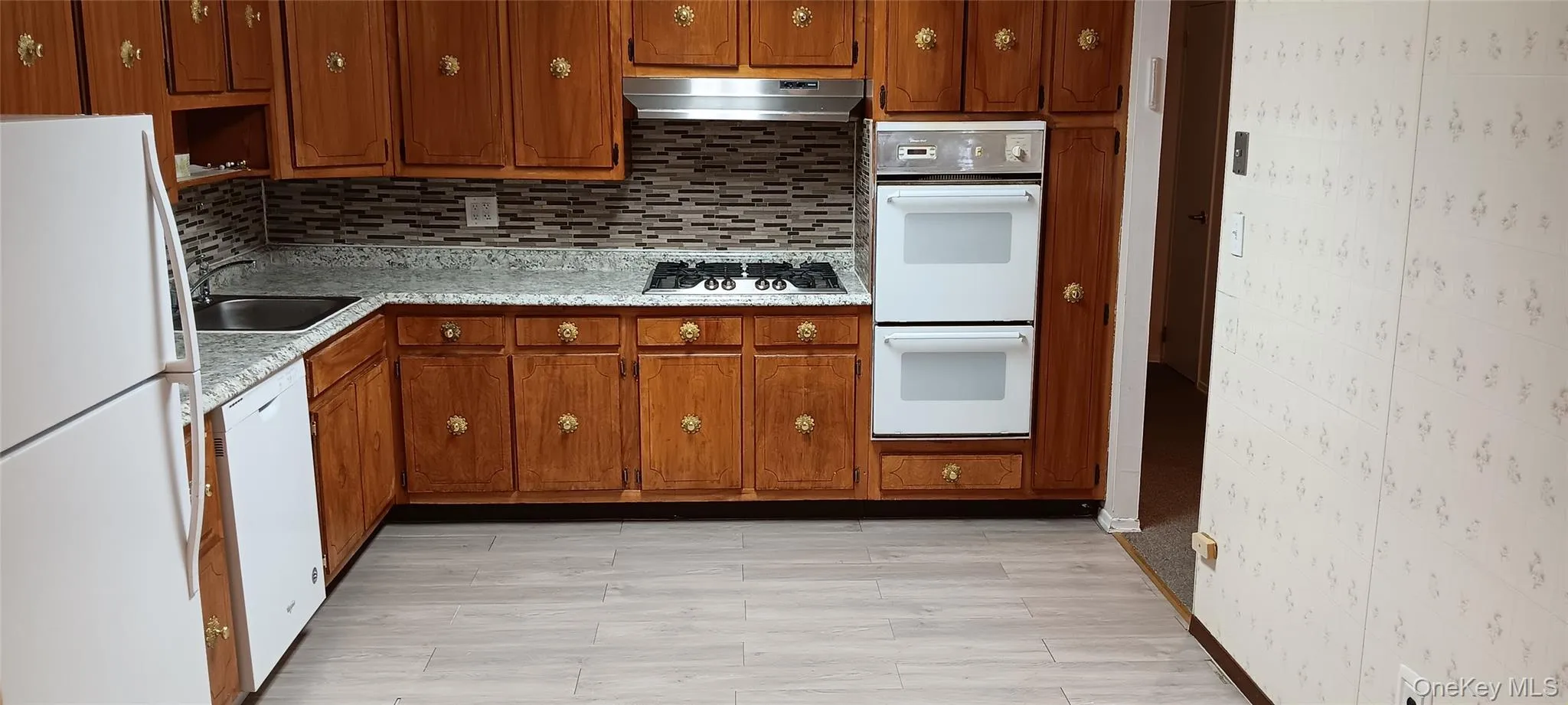 Kitchen with white appliances, light countertops, decorative backsplash, brown cabinets, and under cabinet range hood Kitchen with white appliances, light countertops, decorative backsplash, brown cabinets, and under cabinet range hood