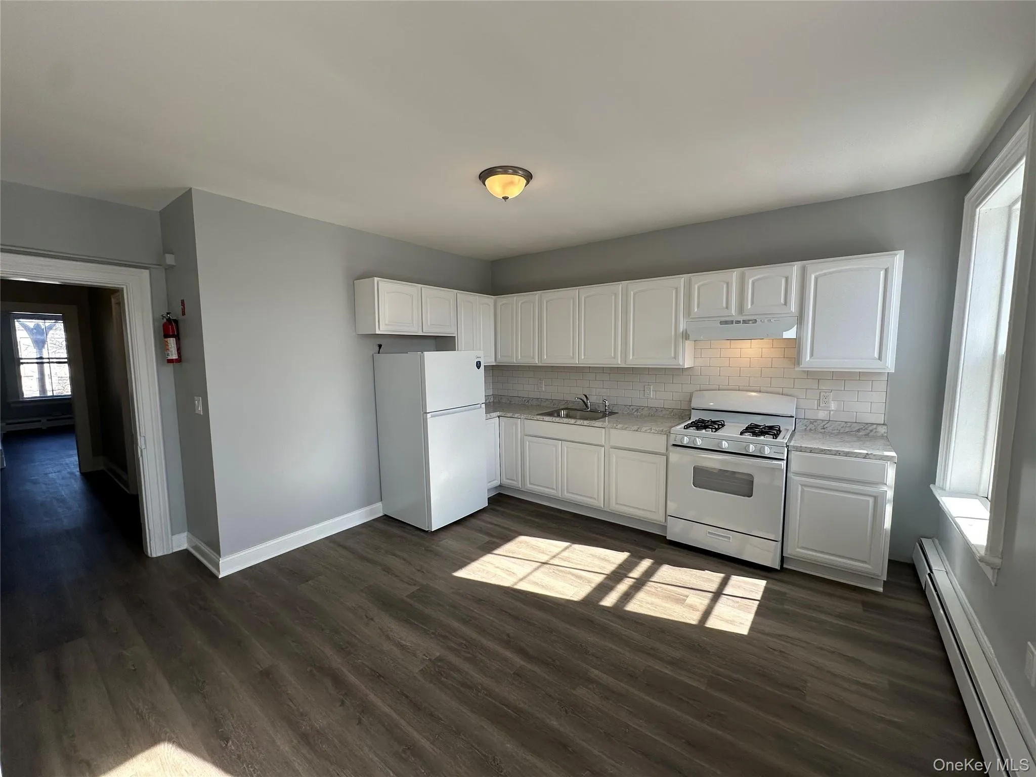 Kitchen with white cabinetry, dark hardwood / wood-style flooring, sink, white appliances, and a baseboard radiator Kitchen with white cabinetry, dark hardwood / wood-style flooring, sink, white appliances, and a baseboard radiator