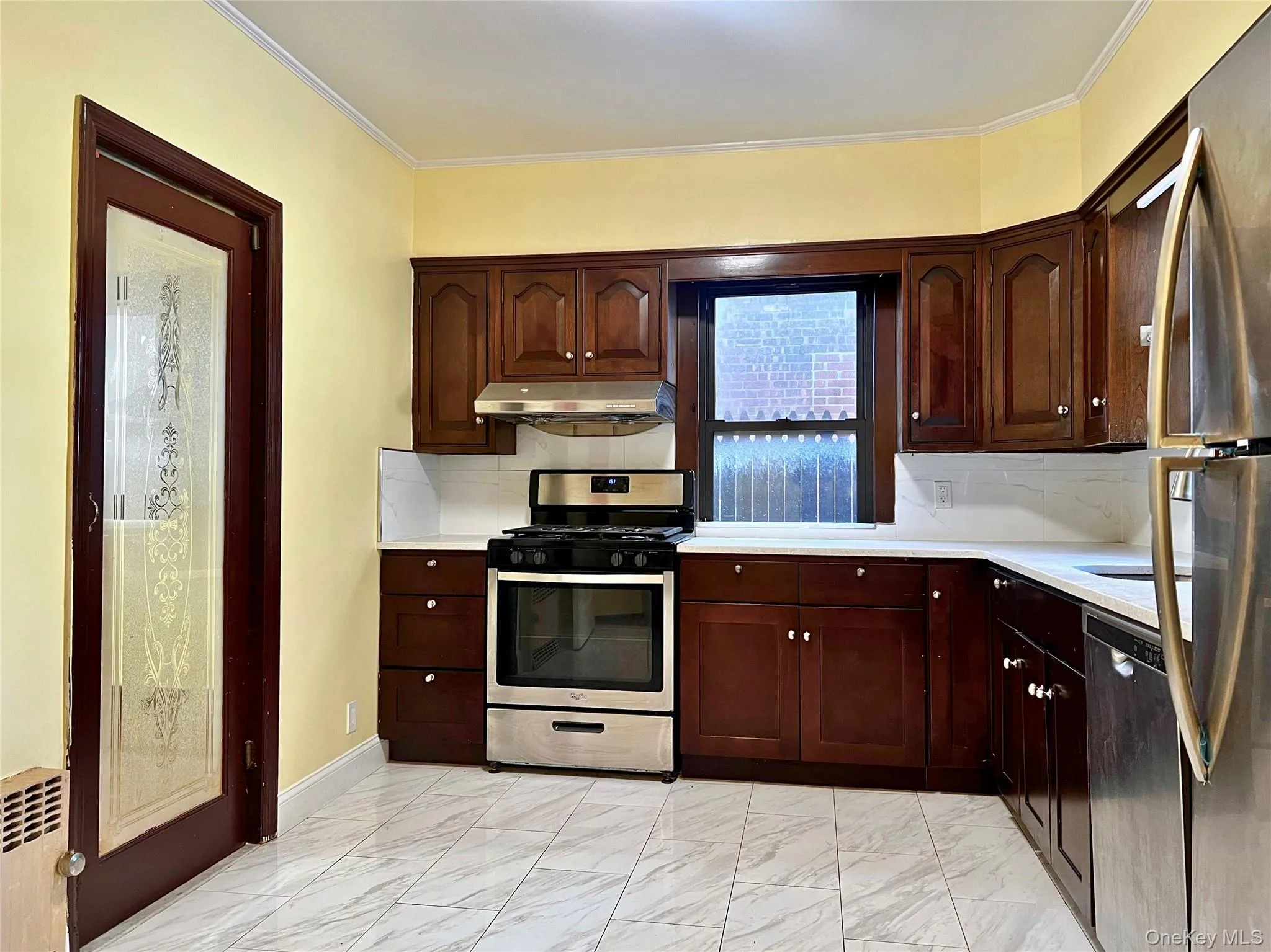 Kitchen with stainless steel appliances, tasteful backsplash, radiator, dark brown cabinetry, and under cabinet range hood Kitchen with stainless steel appliances, tasteful backsplash, radiator, dark brown cabinetry, and under cabinet range hood