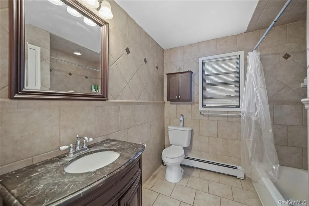 Full bathroom with baseboard heating, shower / tub combo with curtain, vanity, light tile patterned floors, and tile walls Full bathroom with baseboard heating, shower / tub combo with curtain, vanity, light tile patterned floors, and tile walls