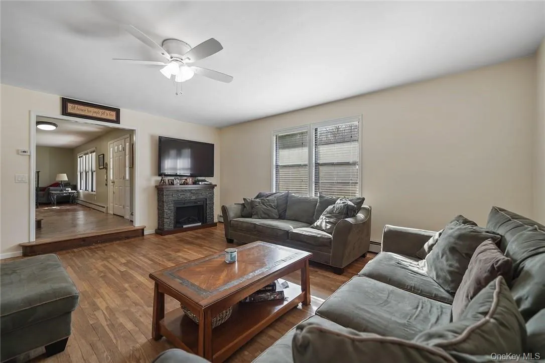 Living room featuring wood finished floors, a stone fireplace, a ceiling fan, and baseboard heating Living room featuring wood finished floors, a stone fireplace, a ceiling fan, and baseboard heating