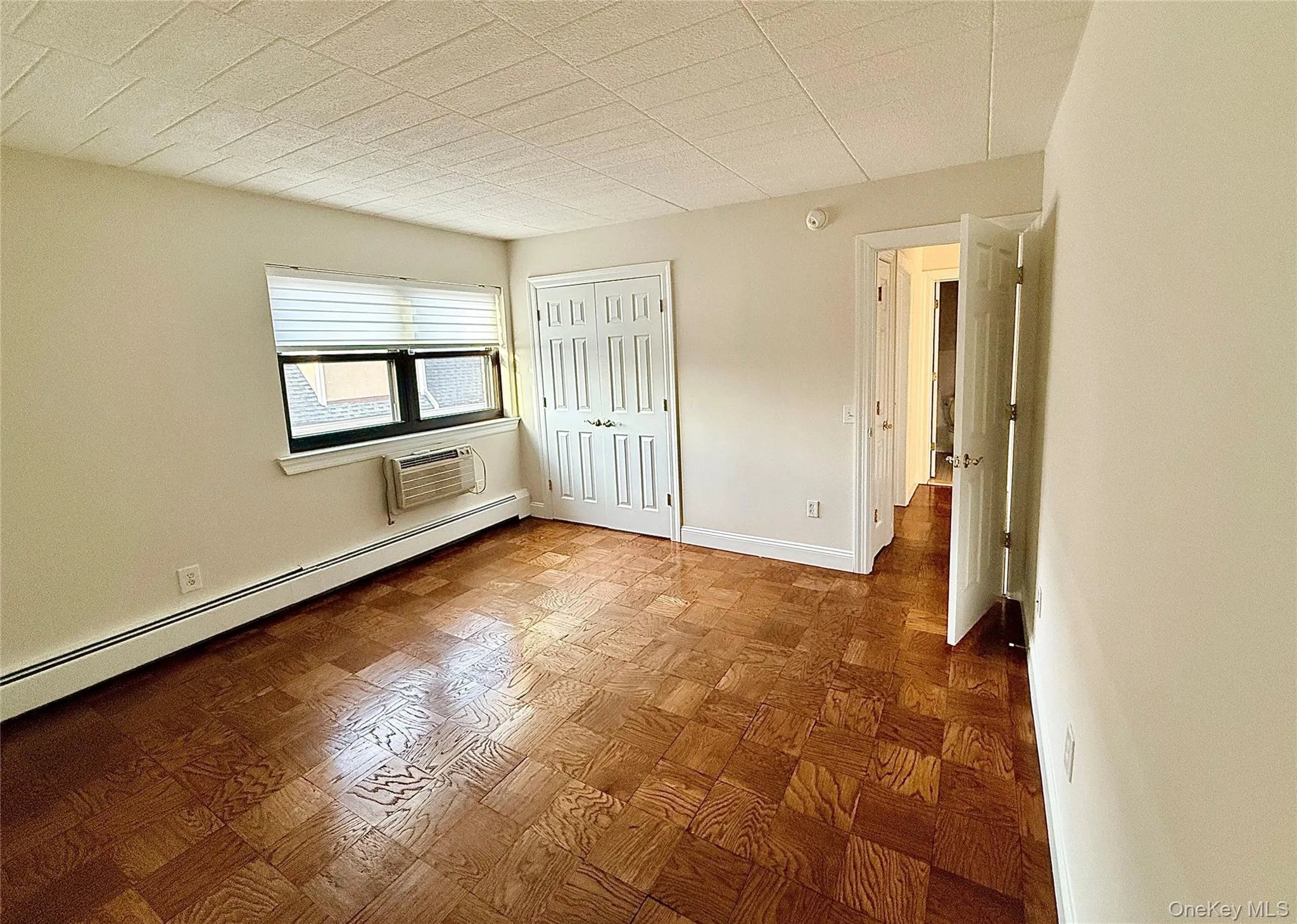 Primary bedroom featuring baseboard heating, a closet, parquet flooring, and an AC wall unit. Primary bedroom featuring baseboard heating, a closet, parquet flooring, and an AC wall unit.