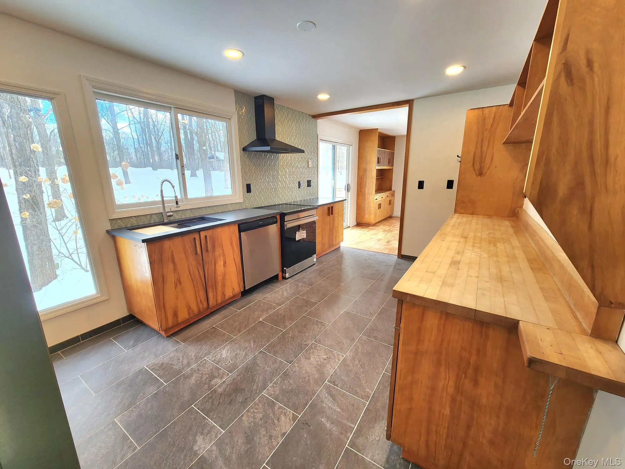 Kitchen with butcher block counters, wall chimney exhaust hood, brown cabinetry, dishwasher, and tasteful backsplash Kitchen with butcher block counters, wall chimney exhaust hood, brown cabinetry, dishwasher, and tasteful backsplash