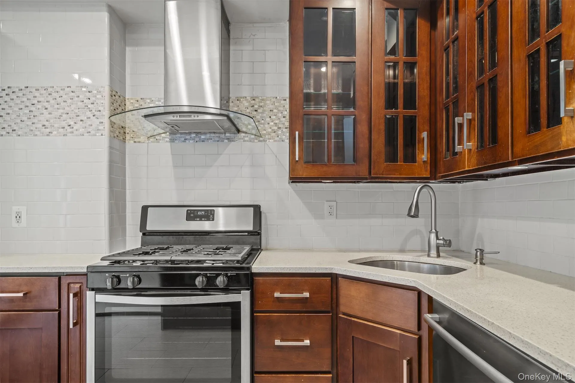 Kitchen with appliances with stainless steel finishes, range hood, backsplash, light stone counters, and glass insert cabinets Kitchen with appliances with stainless steel finishes, range hood, backsplash, light stone counters, and glass insert cabinets