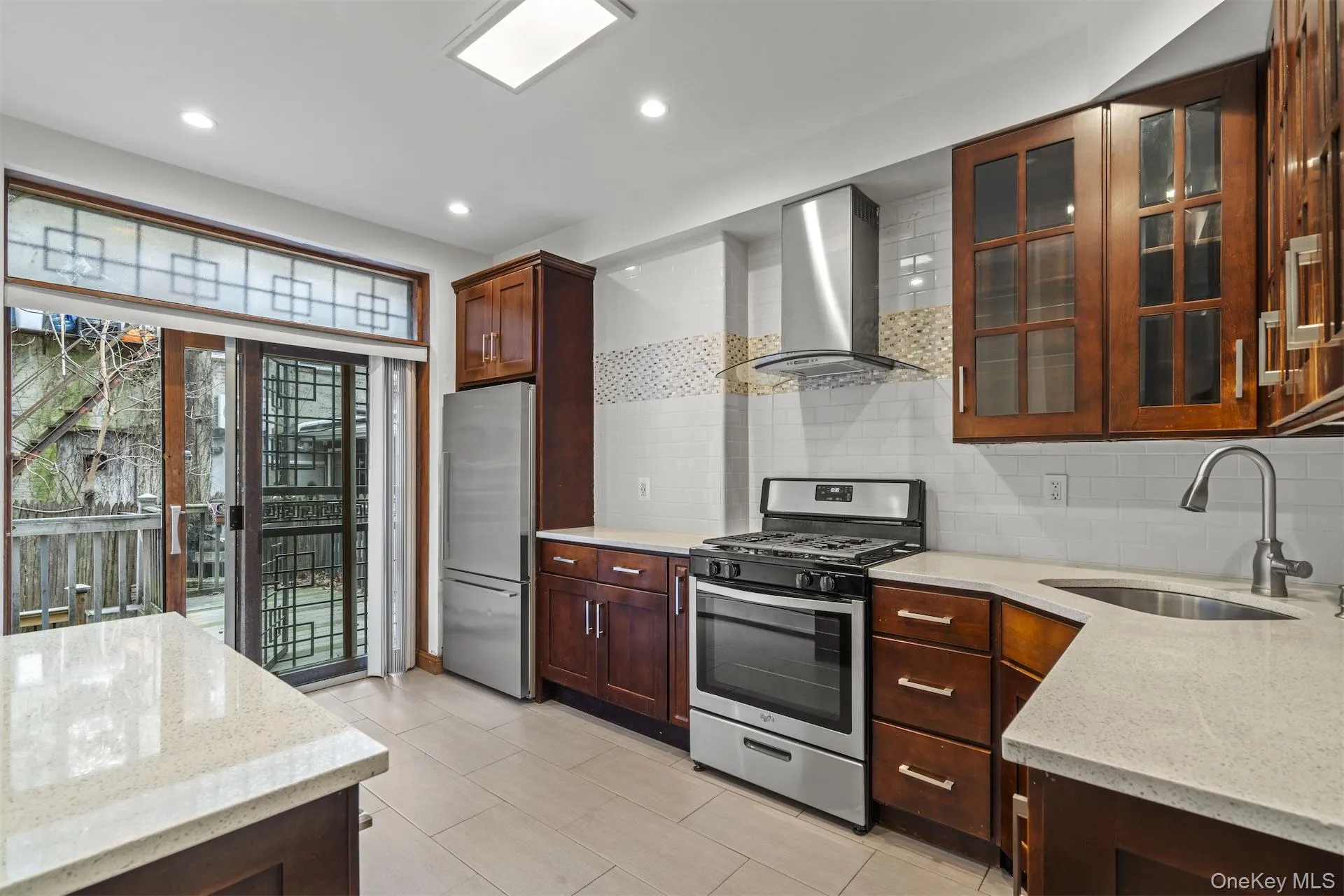 Kitchen with light stone counters, stainless steel appliances, wall chimney range hood, glass insert cabinets, and recessed lighting Kitchen with light stone counters, stainless steel appliances, wall chimney range hood, glass insert cabinets, and recessed lighting