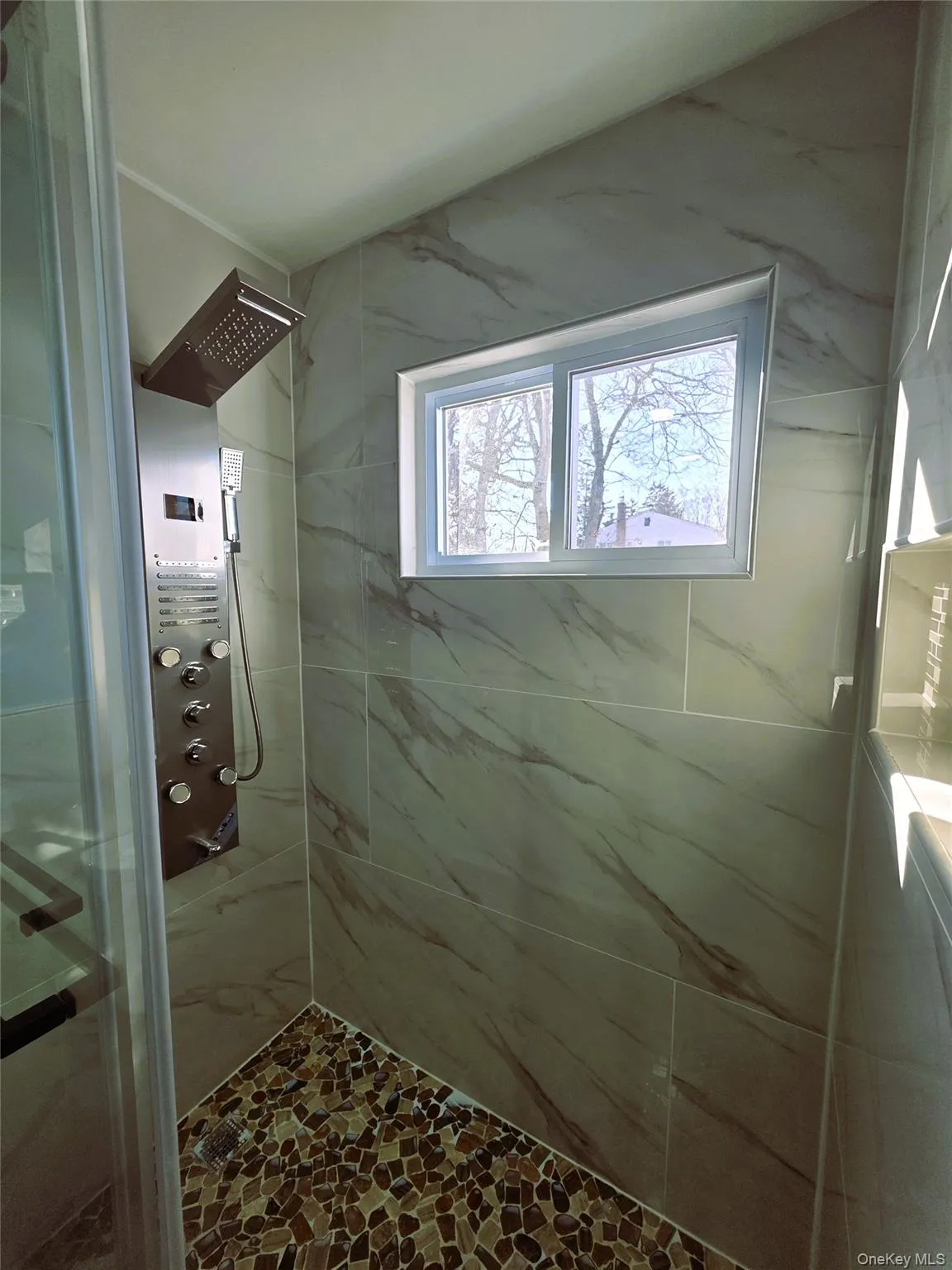 Spacious shower enclosure featuring large format stone-look wall tiles, a pebble-finish shower floor, and a multi-function shower panel Spacious shower enclosure featuring large format stone-look wall tiles, a pebble-finish shower floor, and a multi-function shower panel