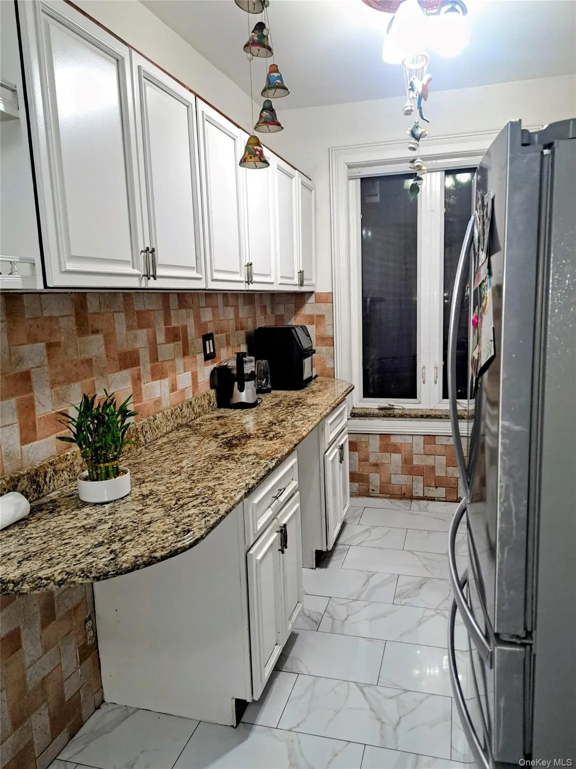 Kitchen with backsplash, freestanding refrigerator, light marble finish flooring, white cabinetry, and light stone counters Kitchen with backsplash, freestanding refrigerator, light marble finish flooring, white cabinetry, and light stone counters