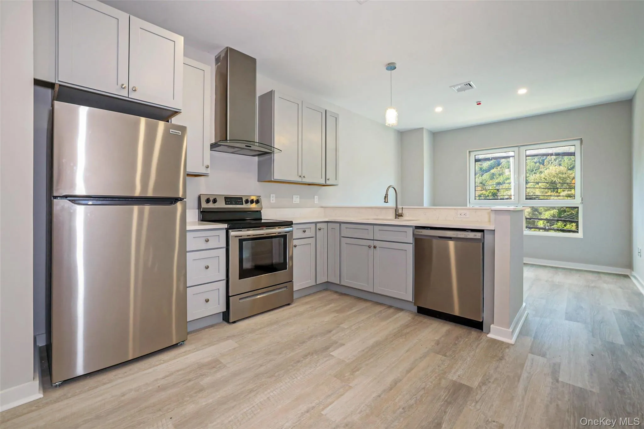 Kitchen featuring appliances with stainless steel finishes, wall chimney range hood, pendant lighting, light wood-style floors, and a peninsula Kitchen featuring appliances with stainless steel finishes, wall chimney range hood, pendant lighting, light wood-style floors, and a peninsula