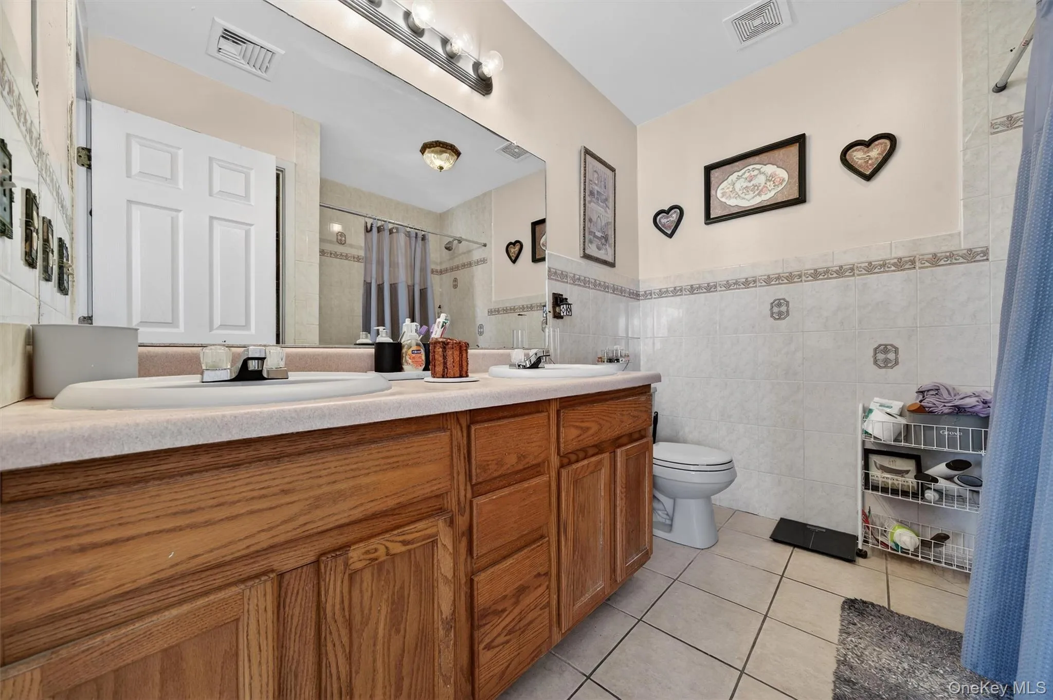Bathroom with light tile patterned flooring, double vanity, tile walls, curtained shower, and wainscoting Bathroom with light tile patterned flooring, double vanity, tile walls, curtained shower, and wainscoting