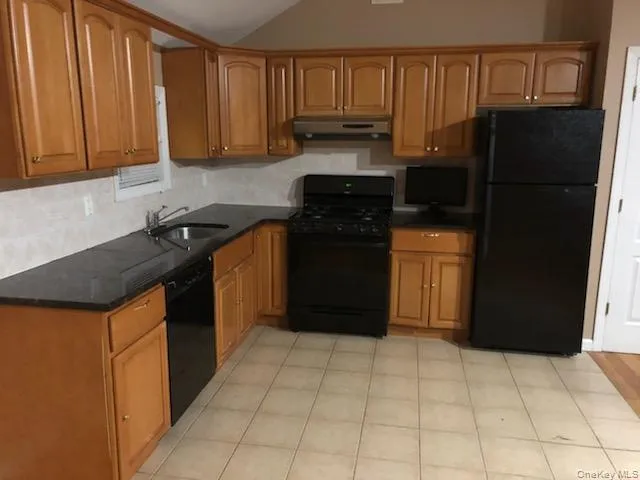 Kitchen featuring black appliances, brown cabinets, under cabinet range hood, lofted ceiling, and tasteful backsplash Kitchen featuring black appliances, brown cabinets, under cabinet range hood, lofted ceiling, and tasteful backsplash