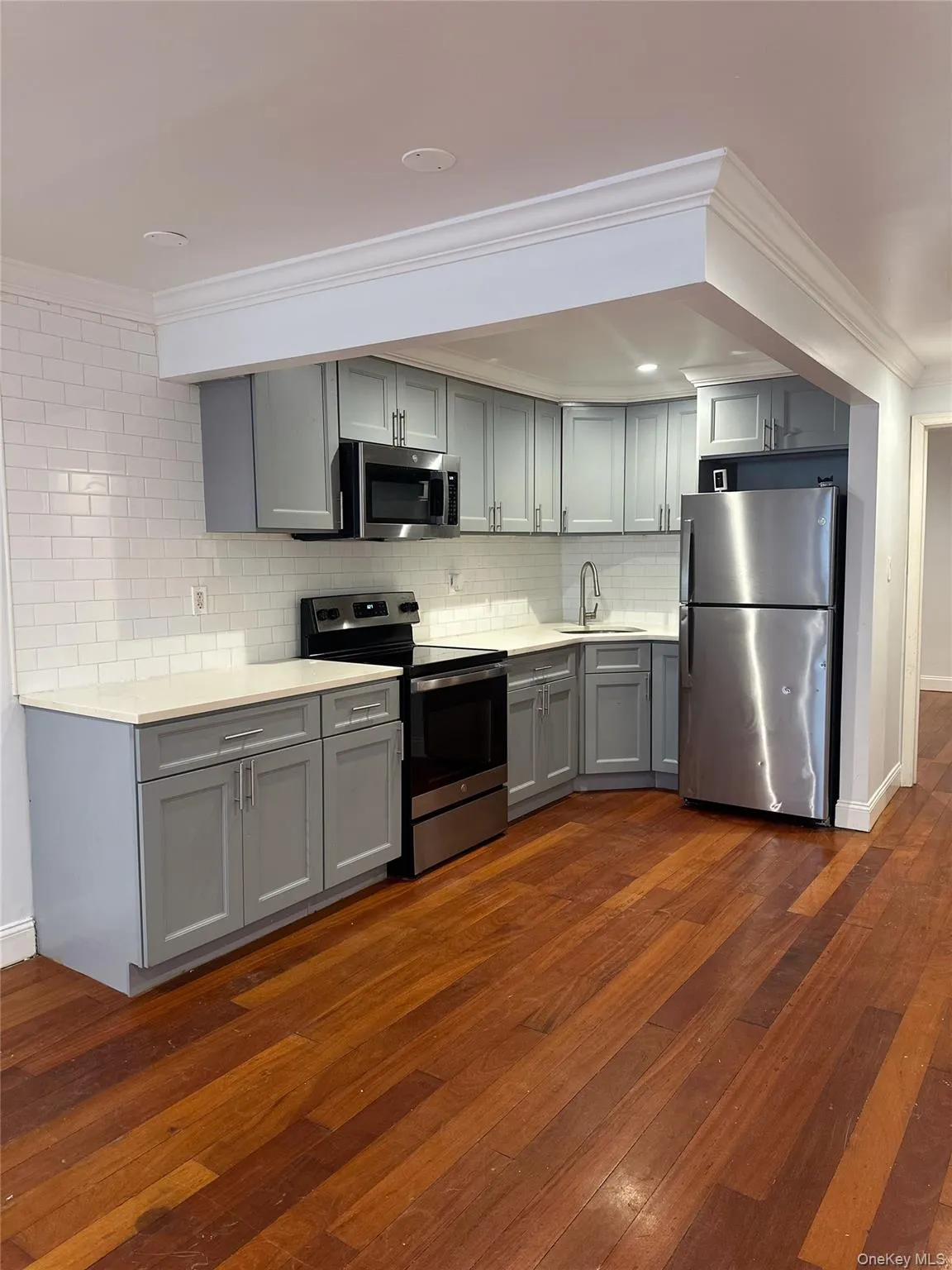 Kitchen with gray cabinetry, ornamental molding, appliances with stainless steel finishes, light countertops, and dark wood-type flooring Kitchen with gray cabinetry, ornamental molding, appliances with stainless steel finishes, light countertops, and dark wood-type flooring