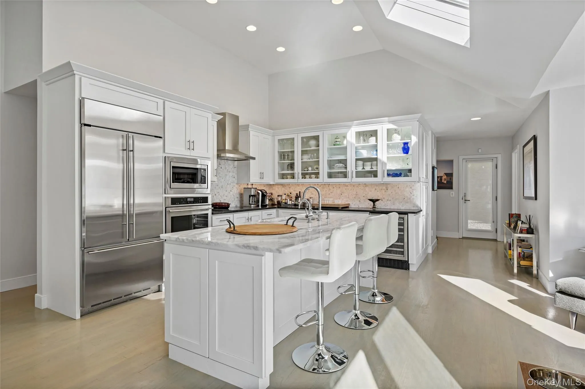 Kitchen with white cabinetry, an island with sink, a breakfast bar area, built in appliances, and wall chimney exhaust hood Kitchen with white cabinetry, an island with sink, a breakfast bar area, built in appliances, and wall chimney exhaust hood