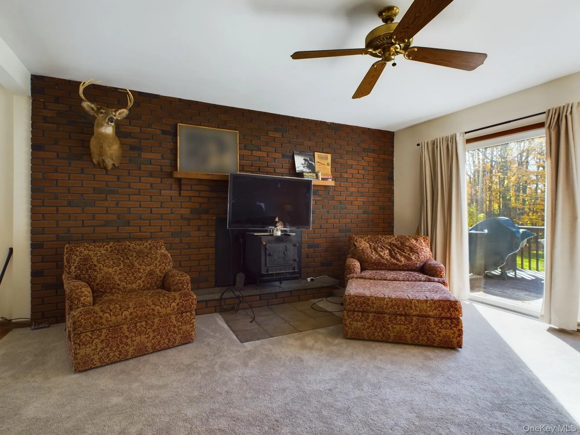 Living room featuring brick wall, carpet floors, a wood stove, and a ceiling fan Living room featuring brick wall, carpet floors, a wood stove, and a ceiling fan