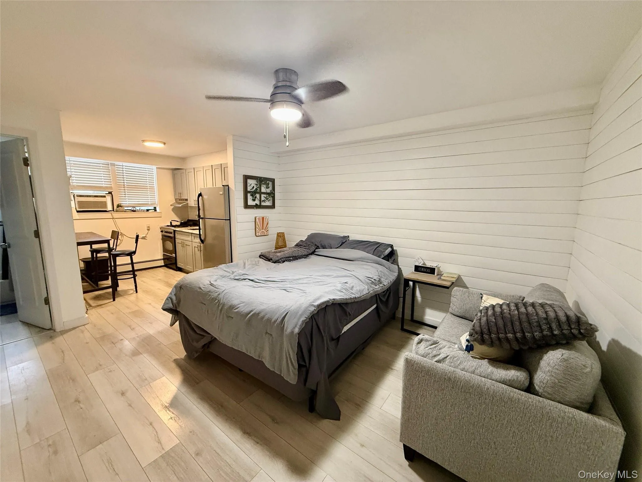 Bedroom with freestanding refrigerator, light wood-style floors, ceiling fan, wood walls, and a baseboard radiator Bedroom with freestanding refrigerator, light wood-style floors, ceiling fan, wood walls, and a baseboard radiator