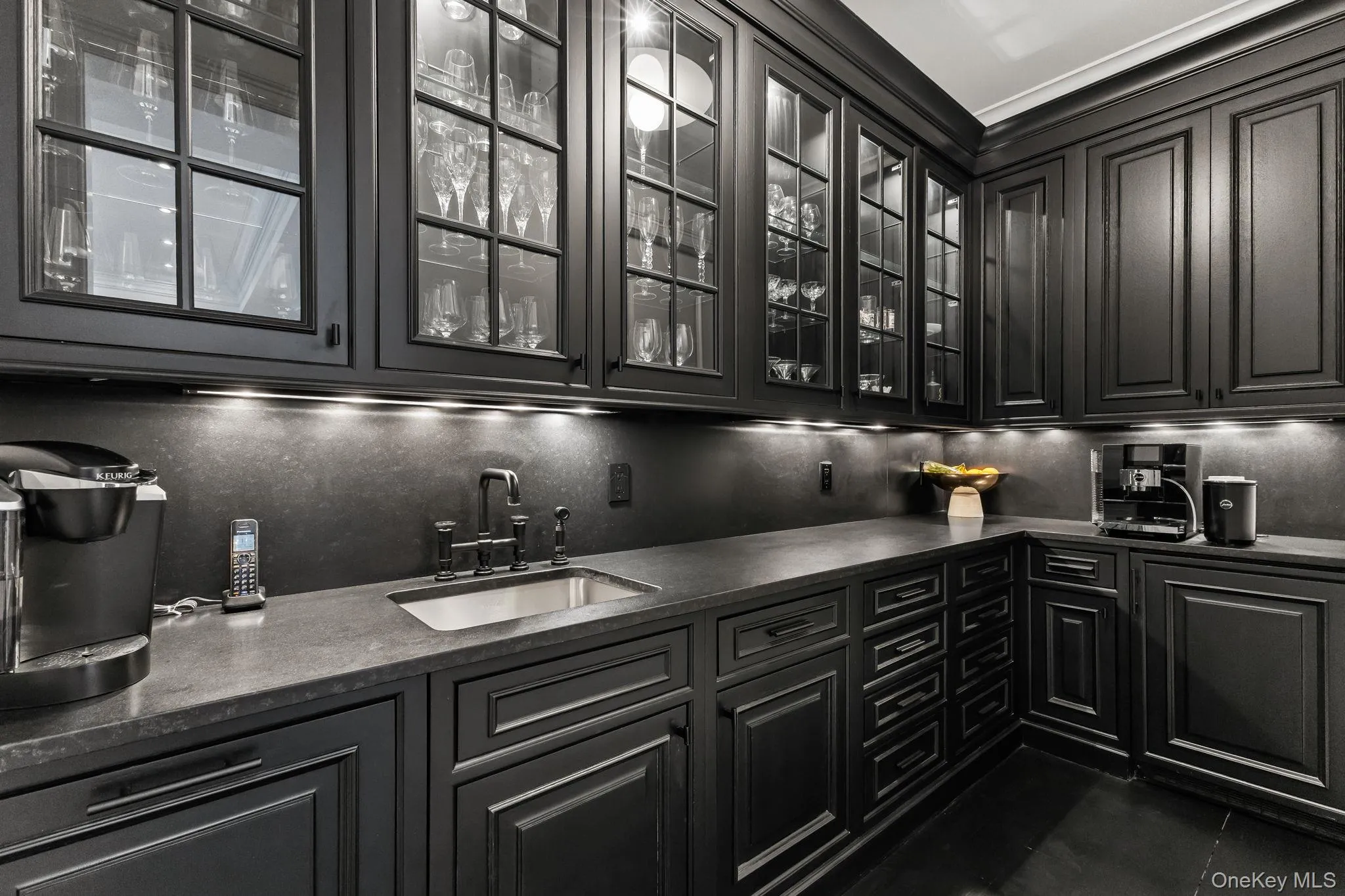 A fully equipped, upscale butler’s pantry featuring custom cabinetry with glass-front upper cabinets and under-cabinet lighting, a stainless steel sink, leathered granite countertops, and bespoke designer lighting. A fully equipped, upscale butler’s pantry featuring custom cabinetry with glass-front upper cabinets and under-cabinet lighting, a stainless steel sink, leathered granite countertops, and bespoke designer lighting.