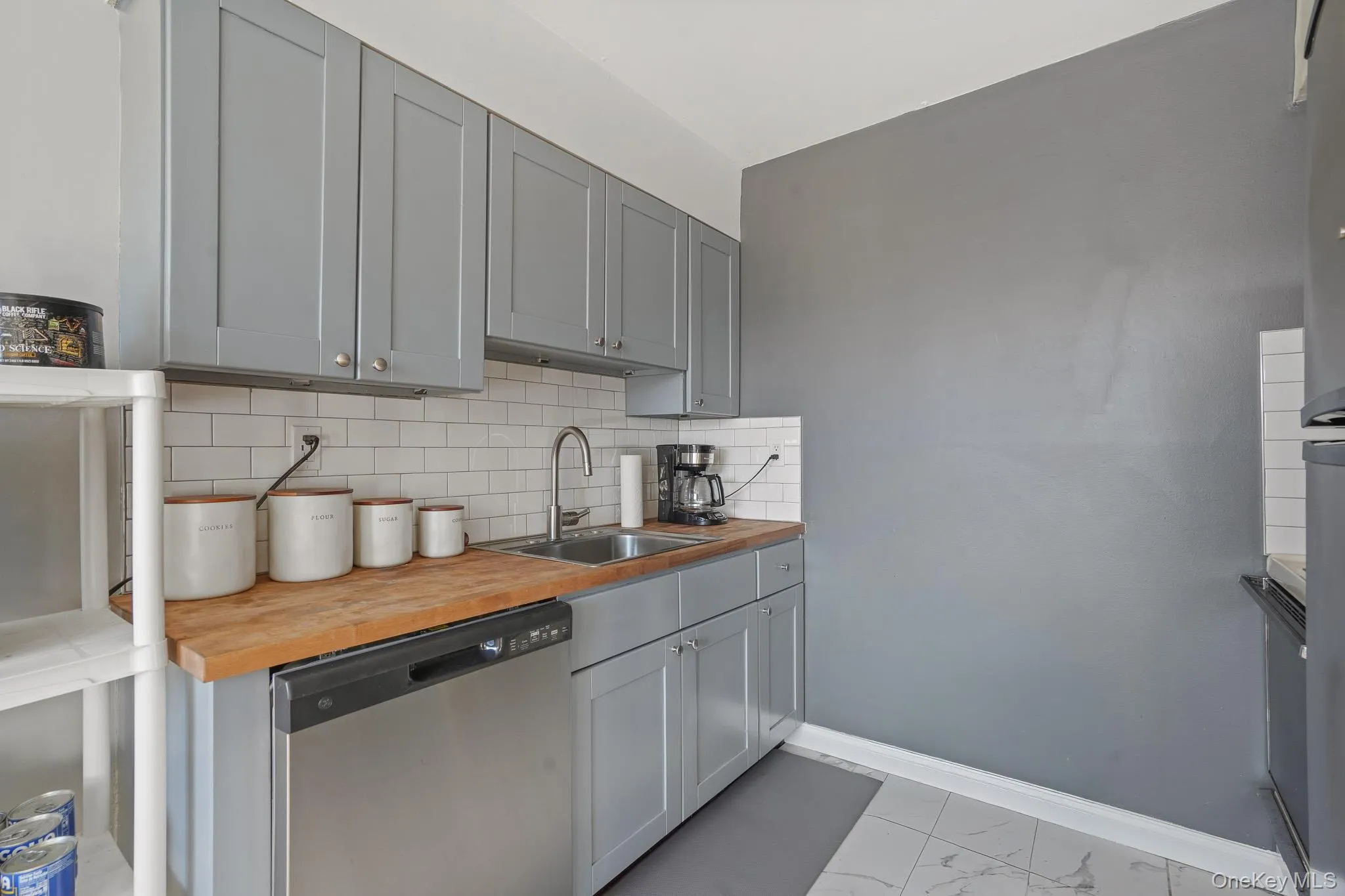 Kitchen featuring gray cabinets, dishwasher, wood counters, backsplash, and light marble finish flooring Kitchen featuring gray cabinets, dishwasher, wood counters, backsplash, and light marble finish flooring
