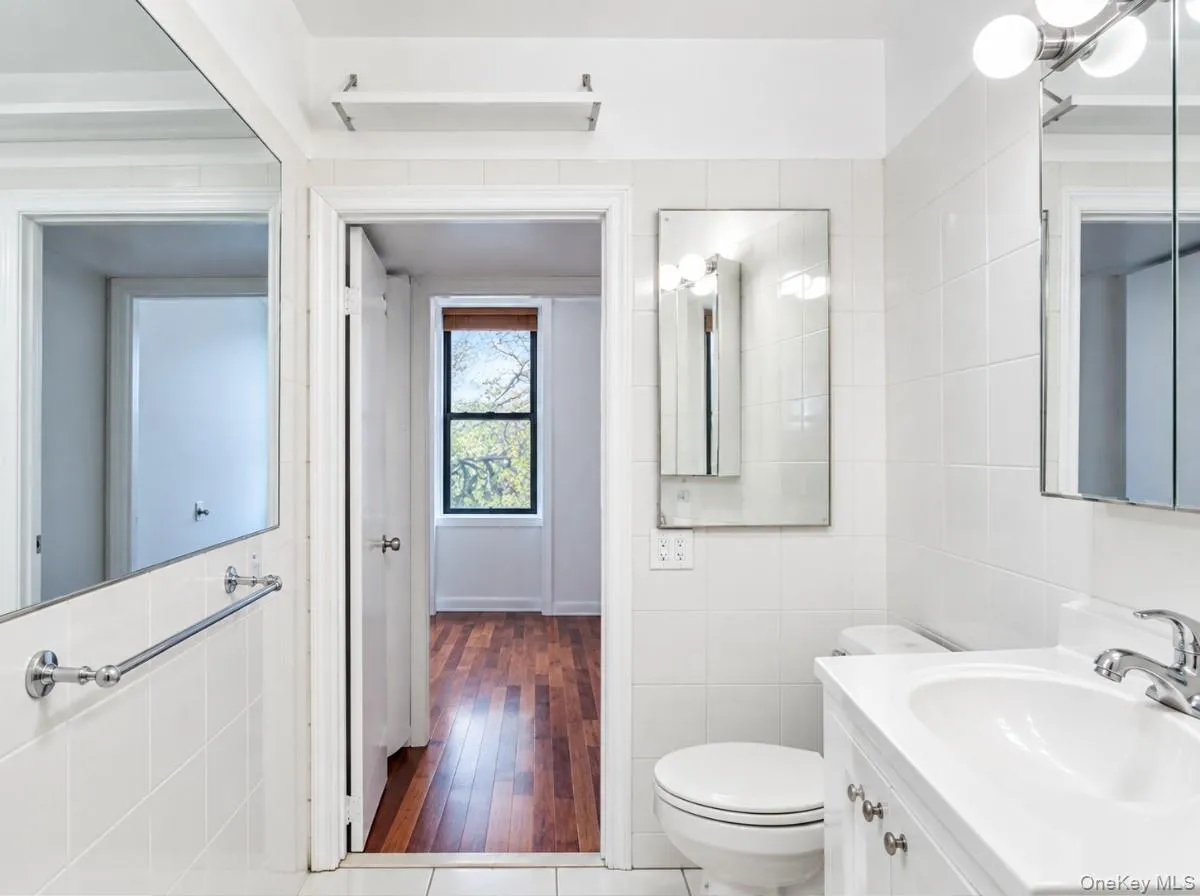 White tiled walls, white vanity with integrated sink, chrome faucet, wall-mounted mirror, and wood-finish flooring visible through the doorway White tiled walls, white vanity with integrated sink, chrome faucet, wall-mounted mirror, and wood-finish flooring visible through the doorway