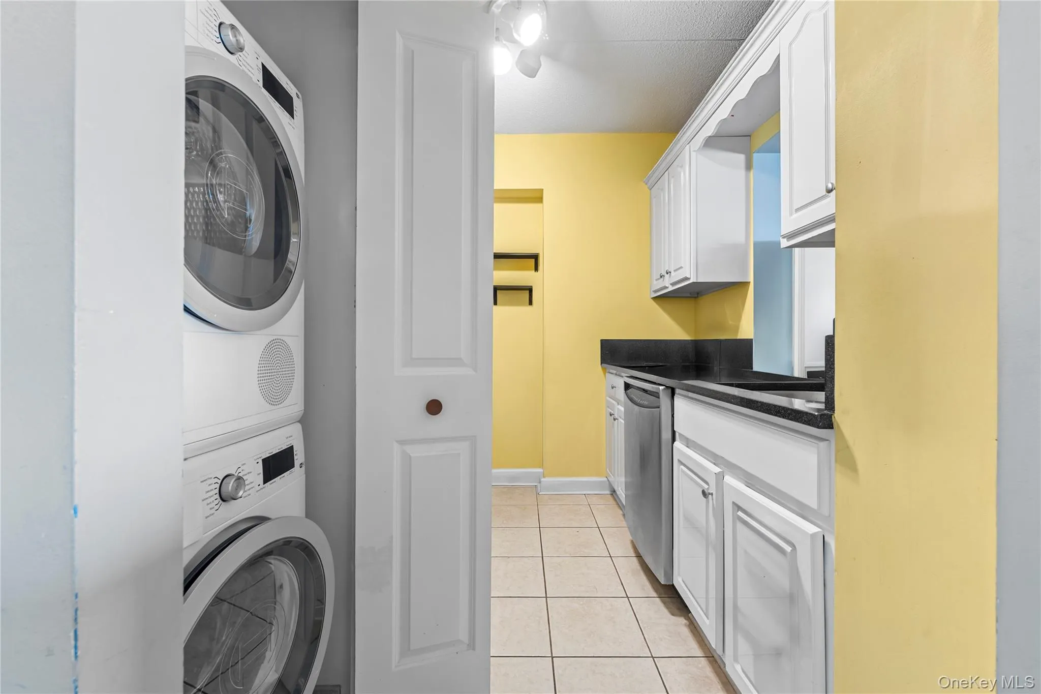 Stackable washer and dryer unit, white cabinetry, black countertops, stainless steel dishwasher, and tile flooring Stackable washer and dryer unit, white cabinetry, black countertops, stainless steel dishwasher, and tile flooring
