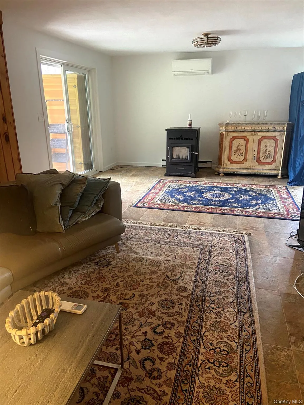 Living room featuring baseboard heating, a wood stove, stone finish floor, and a wall mounted air conditioner Living room featuring baseboard heating, a wood stove, stone finish floor, and a wall mounted air conditioner